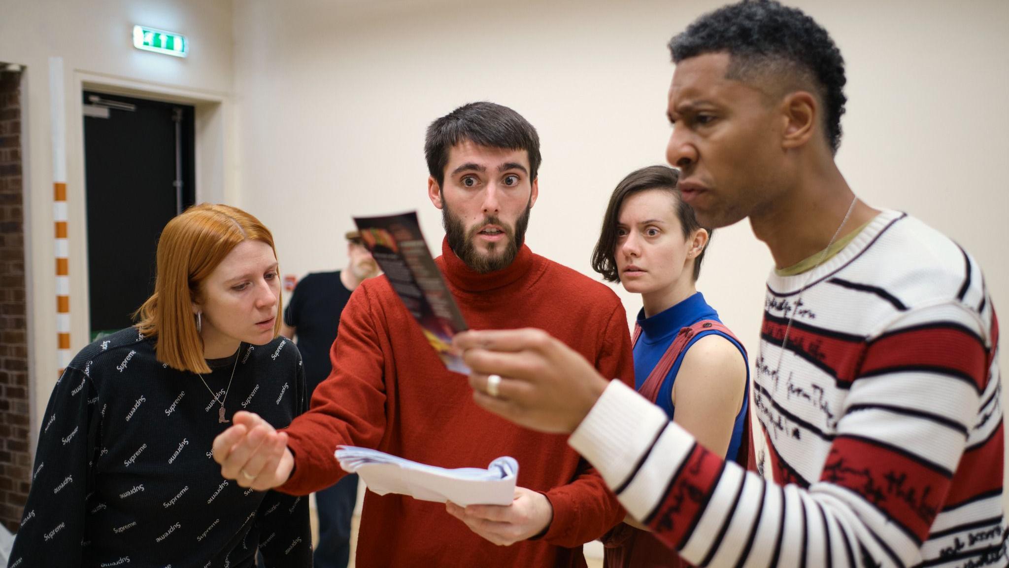 Four actors are standing in a circle in a white room, practising their lines together. They are reading from a script.