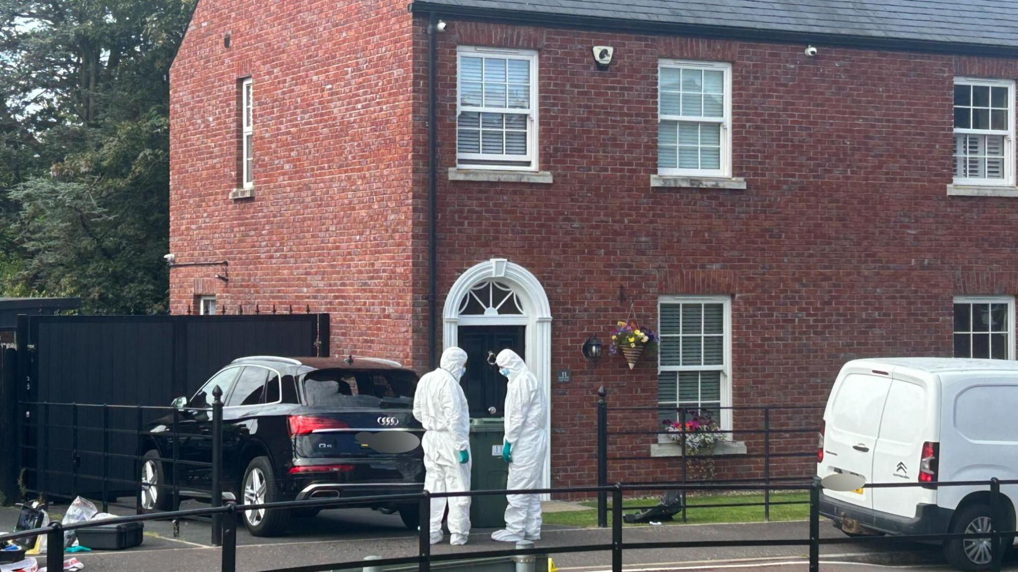 Tow men in police forensic suits stand outside a red brick house. A black car and a white van are parked outside.