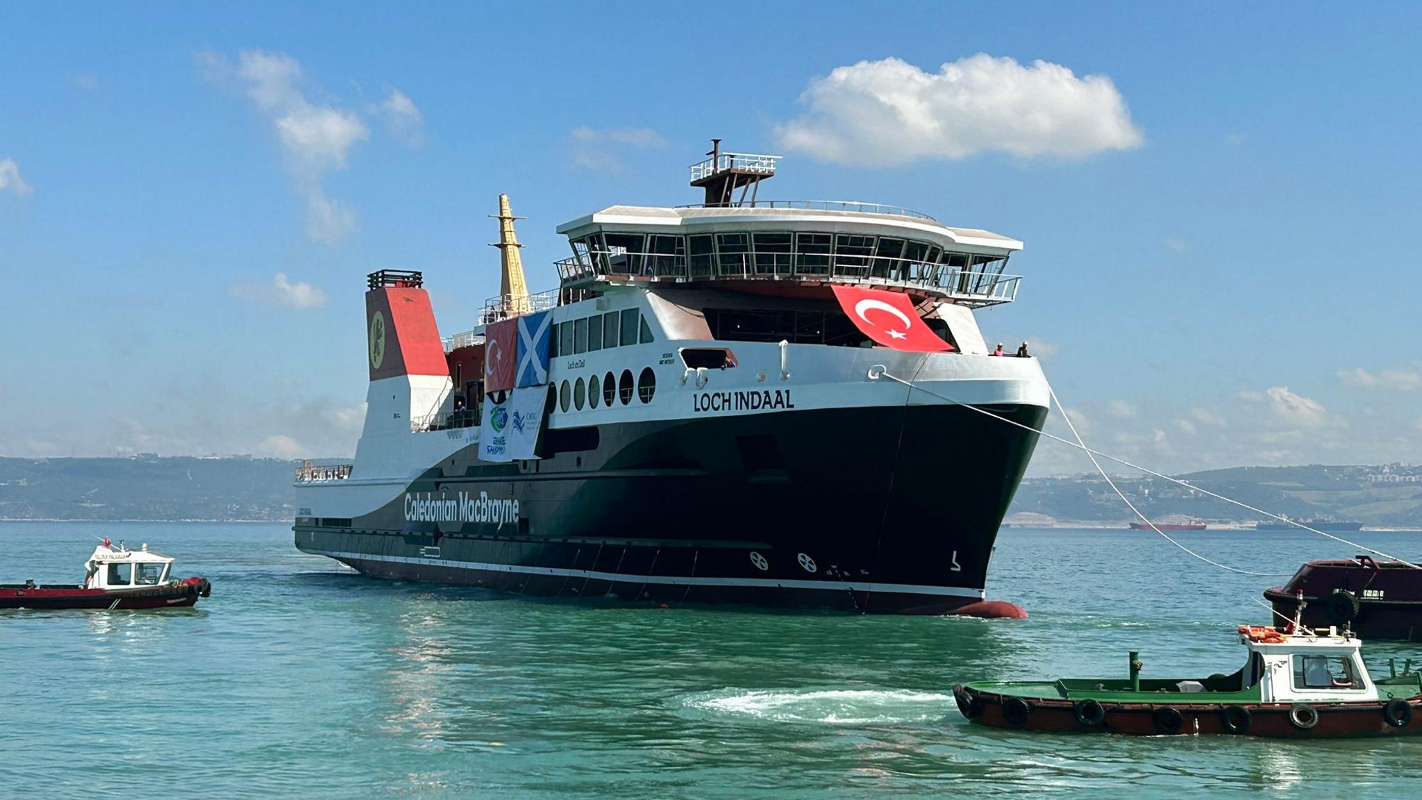 A large black and white ship with red funnels, just launched into the sea with tugs holding it in place