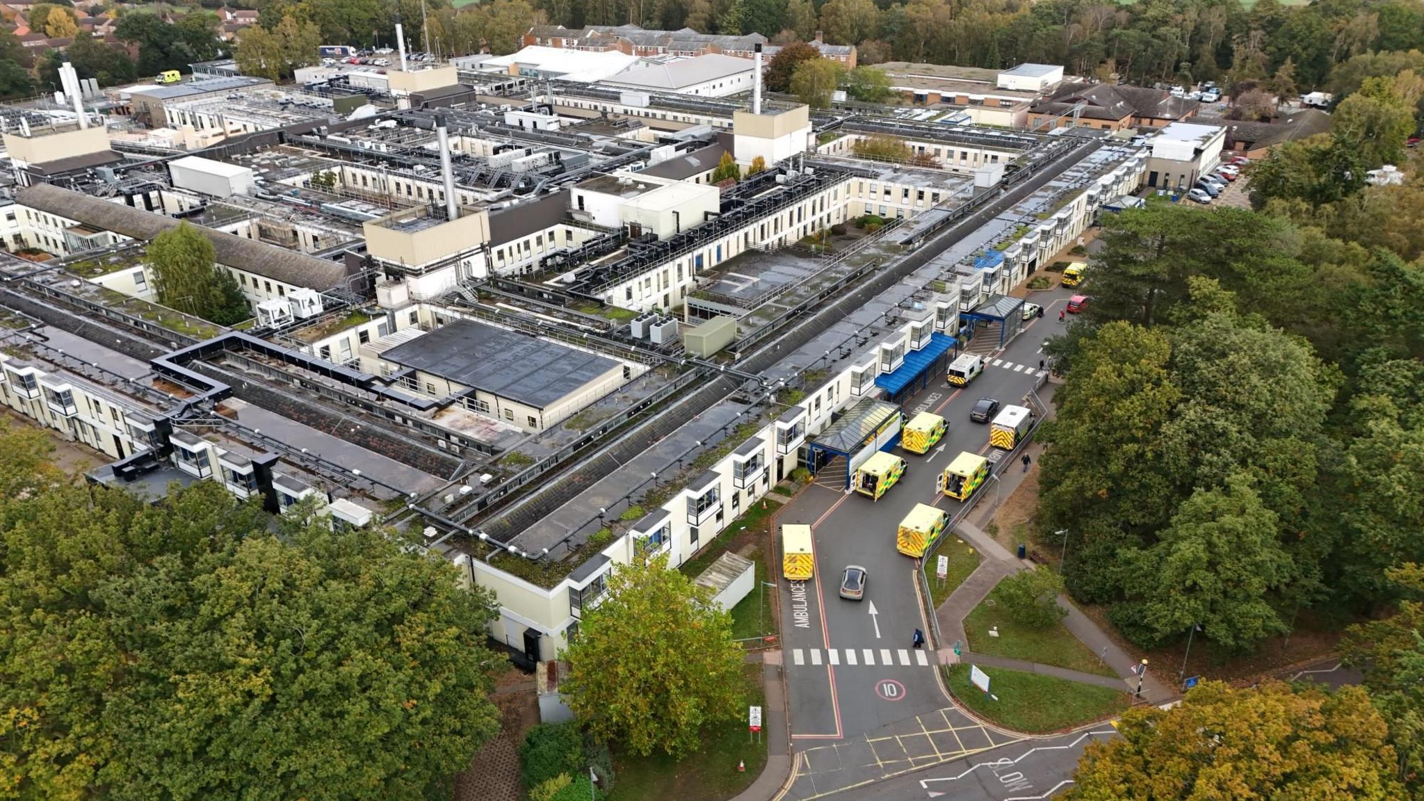 Aerial view of the Queen Elizabeth Hospital in King's Lynn on a cloudy day. It shows the flat, piecemeal makeup of the hospital, which is two storey and has a number of chimneys poking out of the building. The site is sprawling and square and has a light yellow cladded exterior, The photo shows some trees to the south and seven ambulances are queuing outside