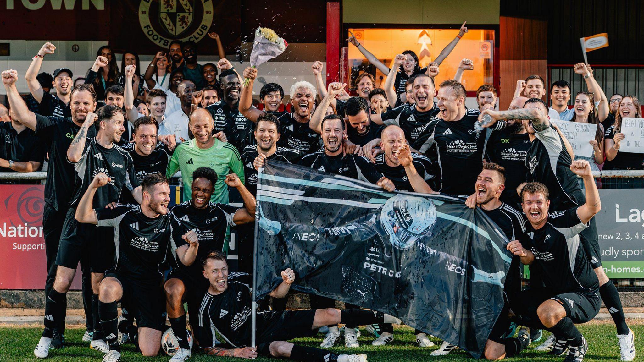 A group of men in black football kit look at the camera and celebrate a win. Some have their hands up in the air. Some are also holding up an image of a Mercedes Formula 1 driver. 