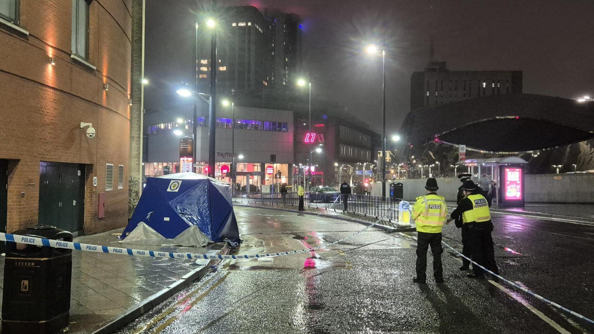 A wet road at night taped off by white and blue police tape. Police officers in yellow hi vis jackets and black hats are standing next to it. There is a blue and white West Midlands police tent on the pavement on the left.