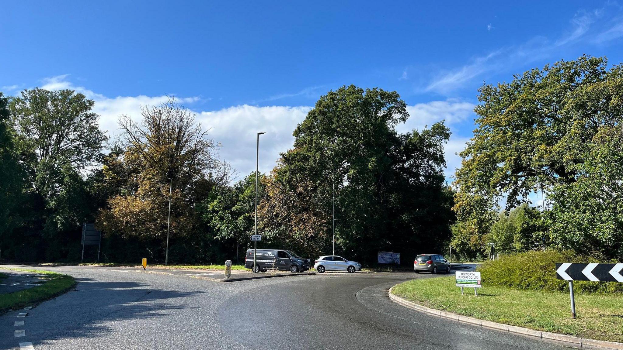 A roundabout seen from the side of the road, with trees along the roadsides and cars driving by.