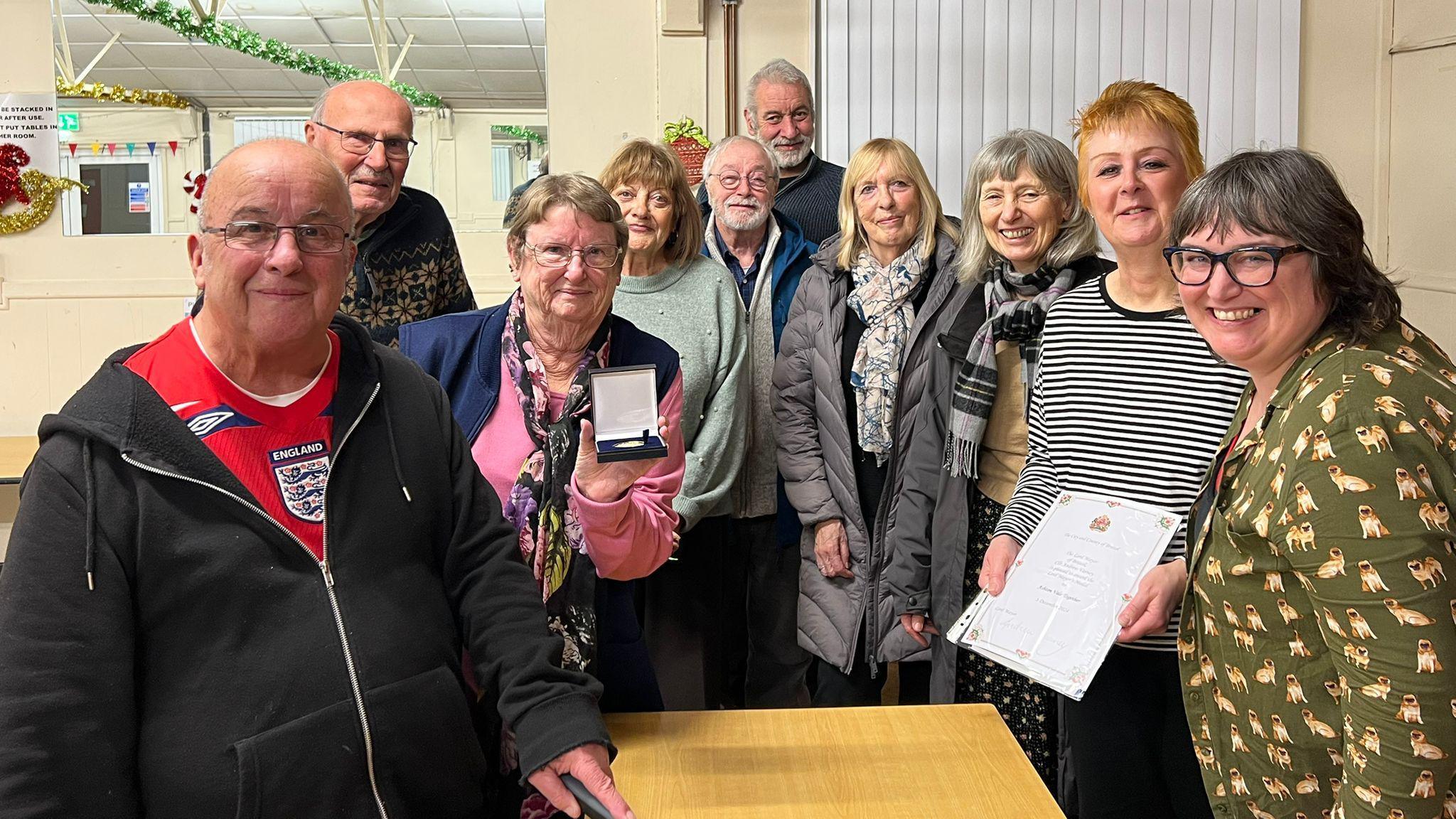 A group of volunteers from Ashton Vale in Bristol stand facing the camera for a group shot. They are a mixture of men and women and are smiling at the camera, with two of them holding up paperwork associated with an award.