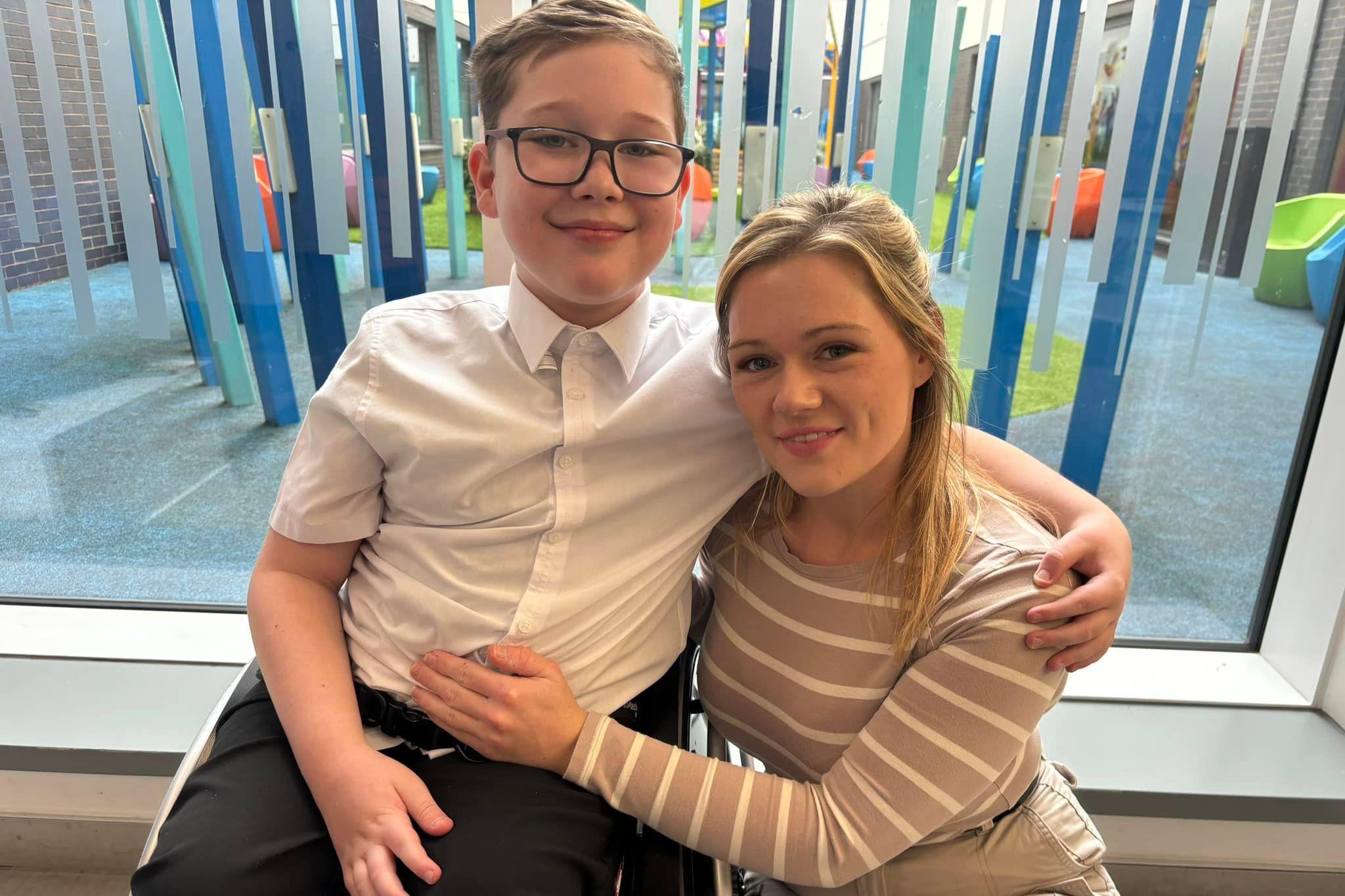 A boy in a white short-sleeved shirt and black trousers sitting in a wheelchair and smiling at the camera, with his arm wrapped around a blonde woman who kneels next to him. She has blonde hair and wears a brown and white stripey top, with her hand on the boy's stomach. They sit in front of a window, which shows a multicoloured sculpture outside.