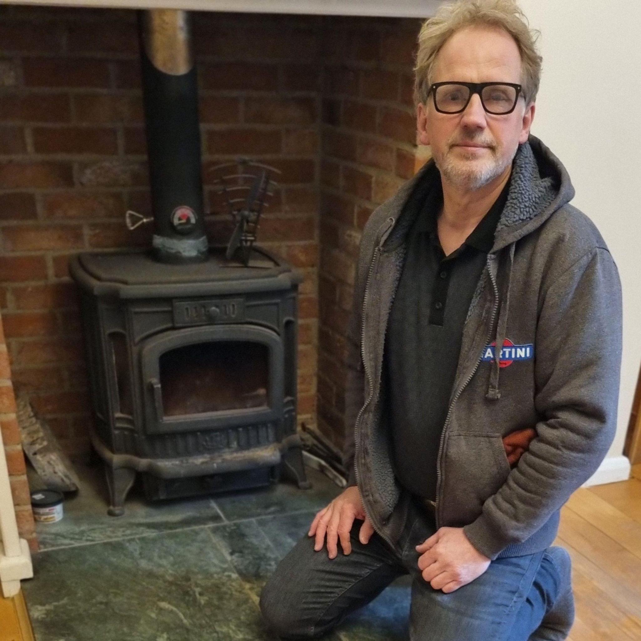 Kerry Walpole is kneeling in front of a wood burner. He's wearing a grey fleece over a dark T-shirt and jeans.