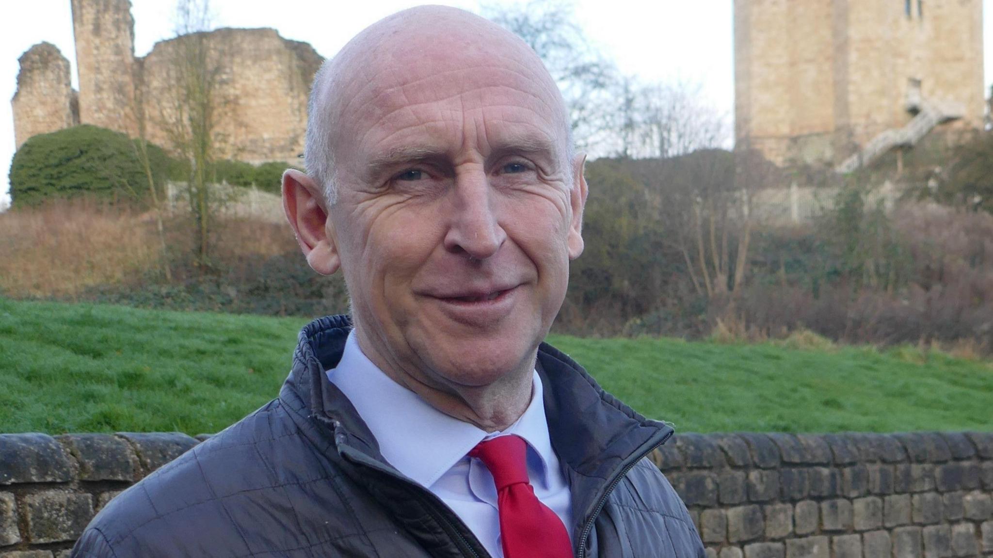 A man with a shaved head and blue eyes wearing a light blue shirt, red tie and navy jacket. He stands in front of a low dark wall. Behind the wall is a grass bank, trees and hedges and some ruined buildings in the distance.