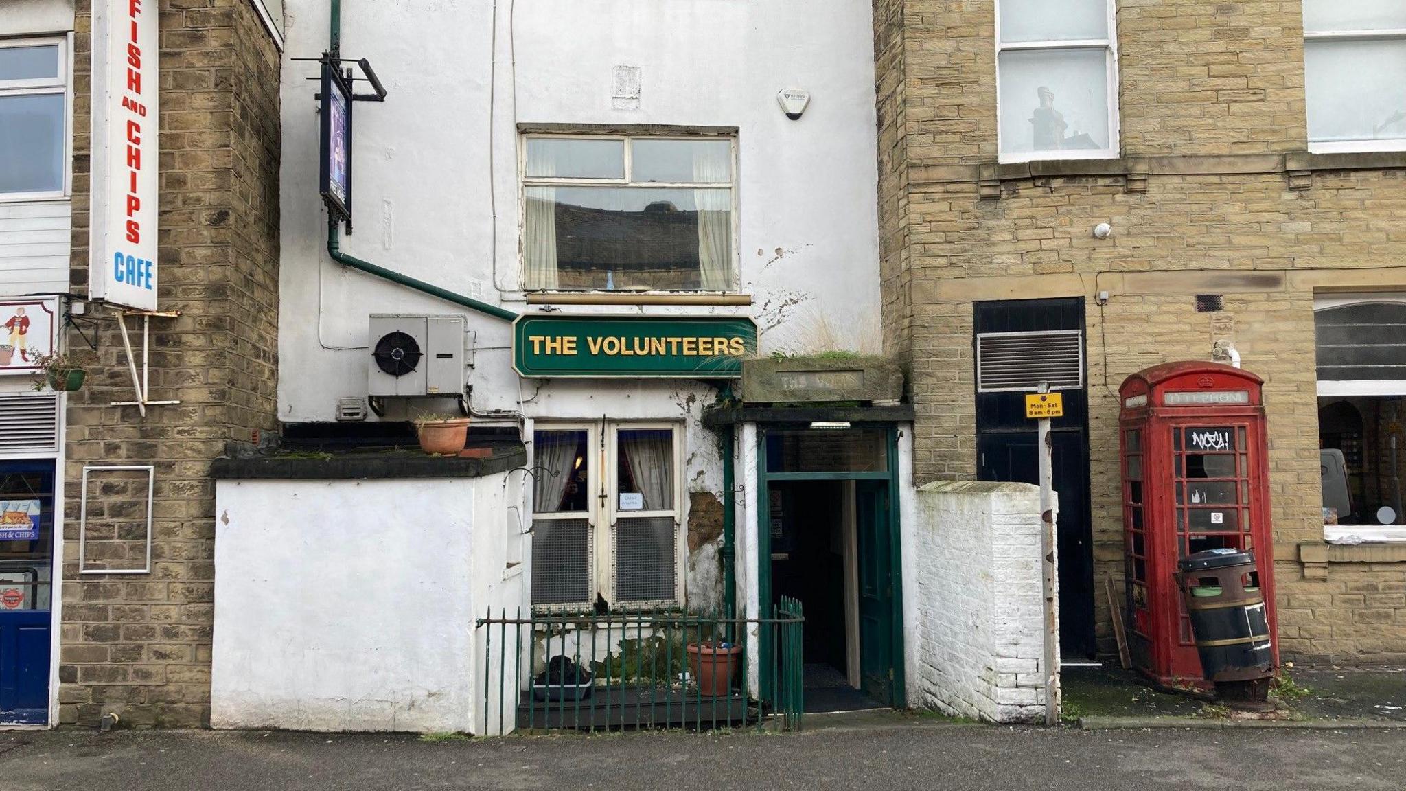 A doorway with green wooden framing into the pub which has crumbling whitewashed walls and a sign in gold lettering with a green background spelling out the word 'The Volunteers'.