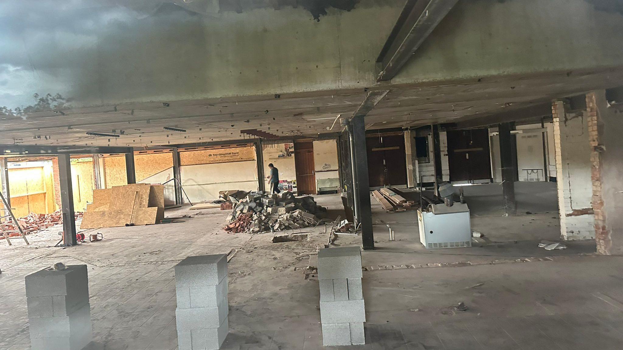 A large empty building with no rooms. There are exposed beams and concrete blocks. There is a pile of rubble in the centre and a person is in the background.