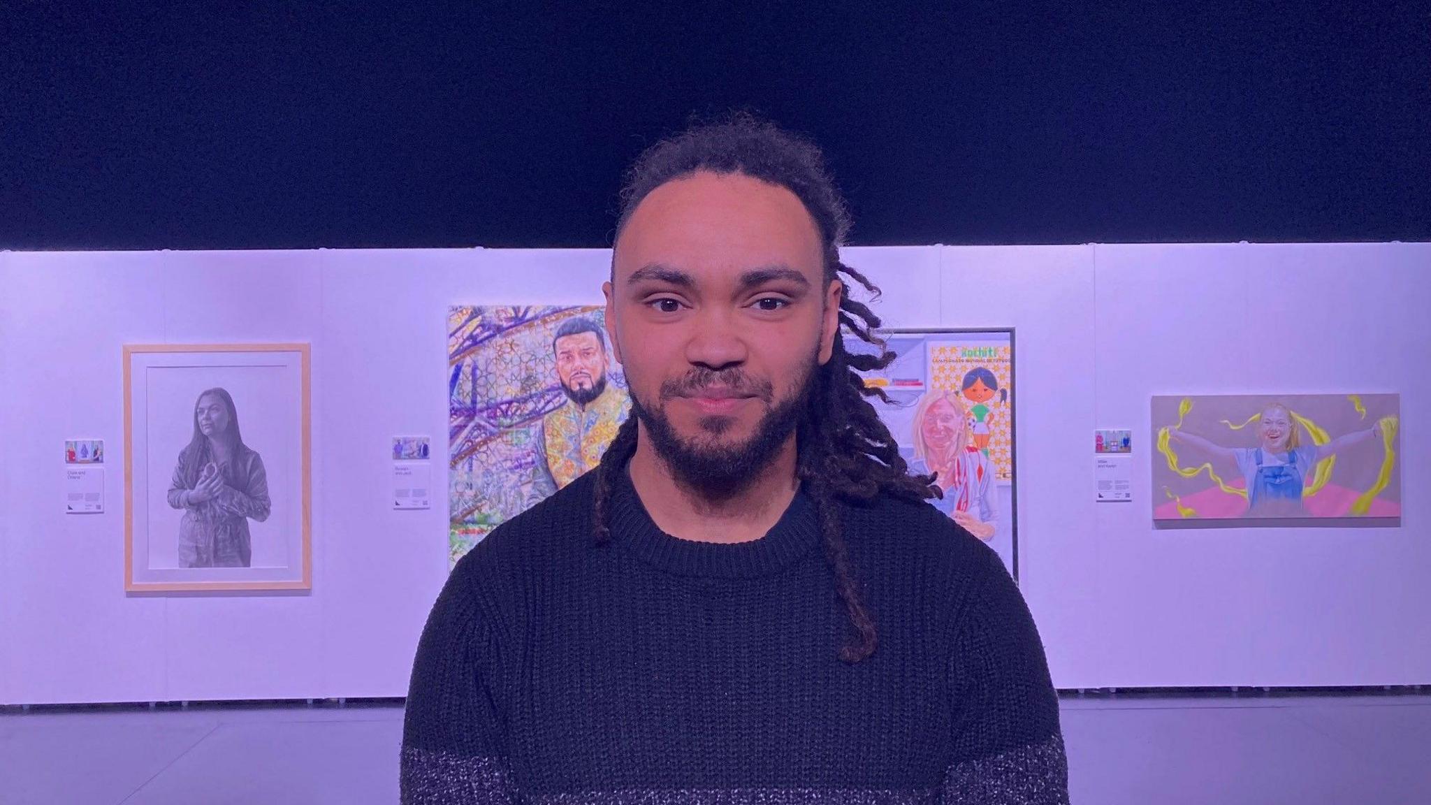 A man with long, tied back dreadlocks and a black jumper with paintings behind him on a white screen. 