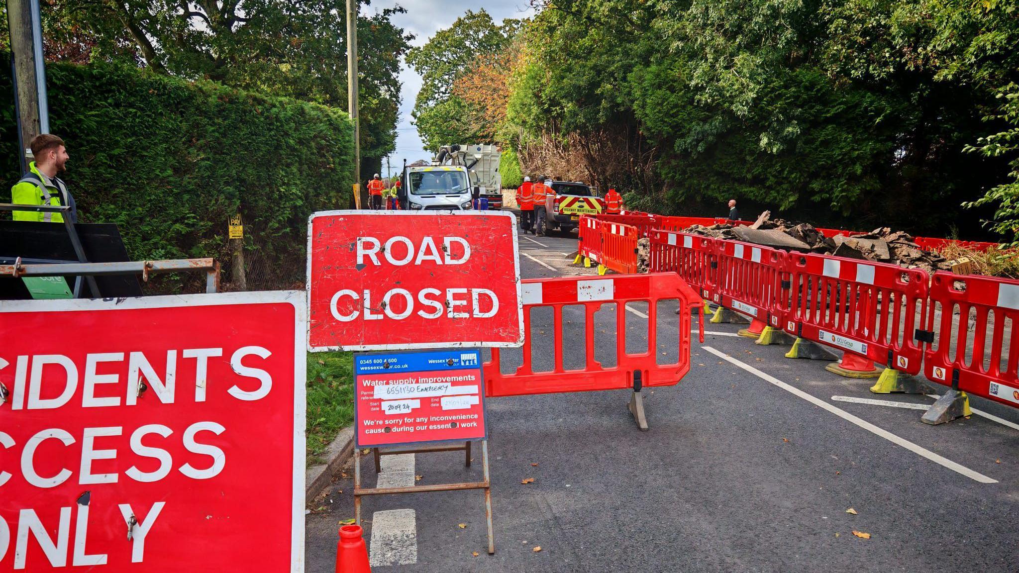 Image shows a road closure sign in the middle of a road, with red fences surrounding a burst pipe. 