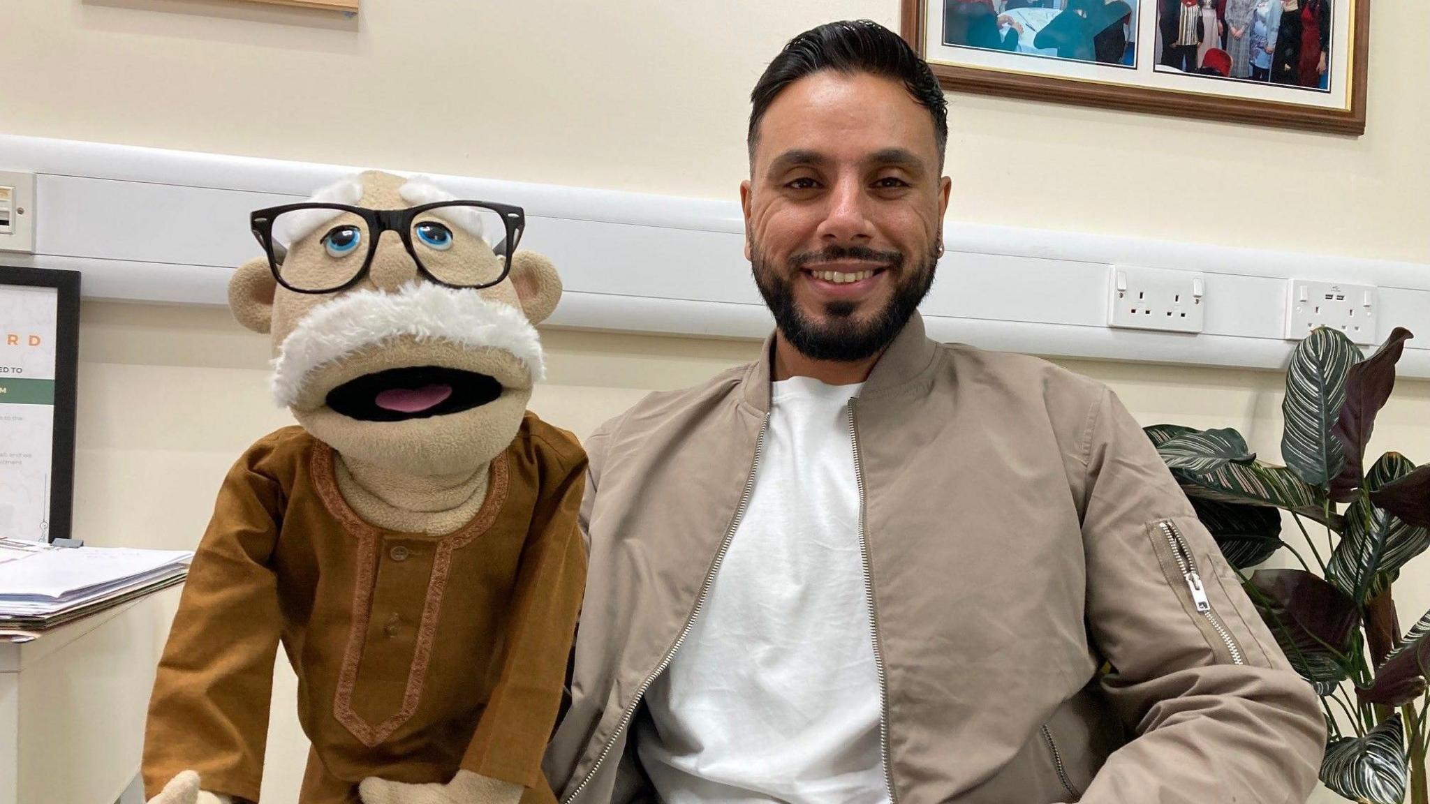 A hand puppet with a white moustache and glasses with the puppeteer who is wearing a light brown jacket and white T-shirt underneath.   