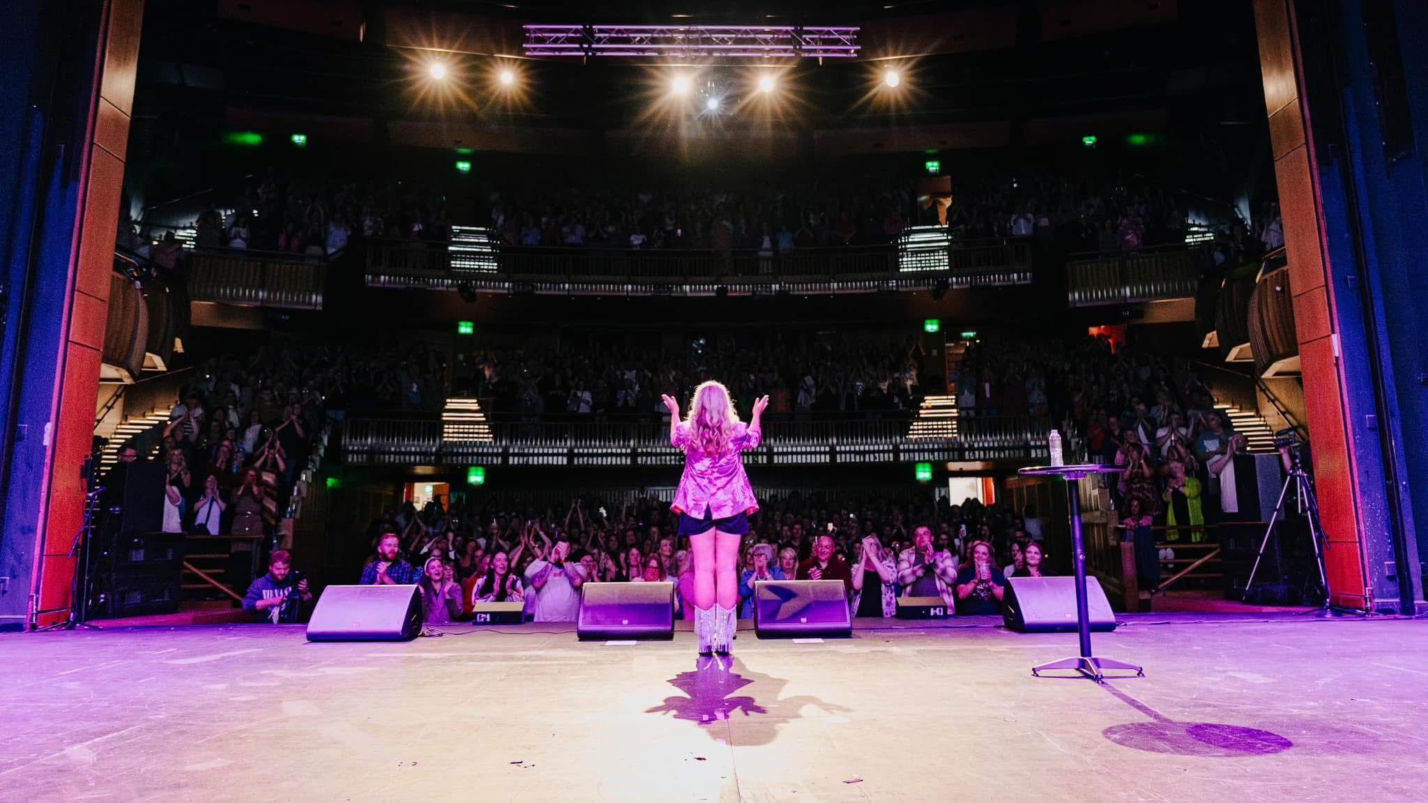 A picture from behind Ms Terry on stage. The audience are facing the camera but are in slight darkness. Ms Terry is facing them, wearing a pink jacket and she is standing in the spotlight. 
