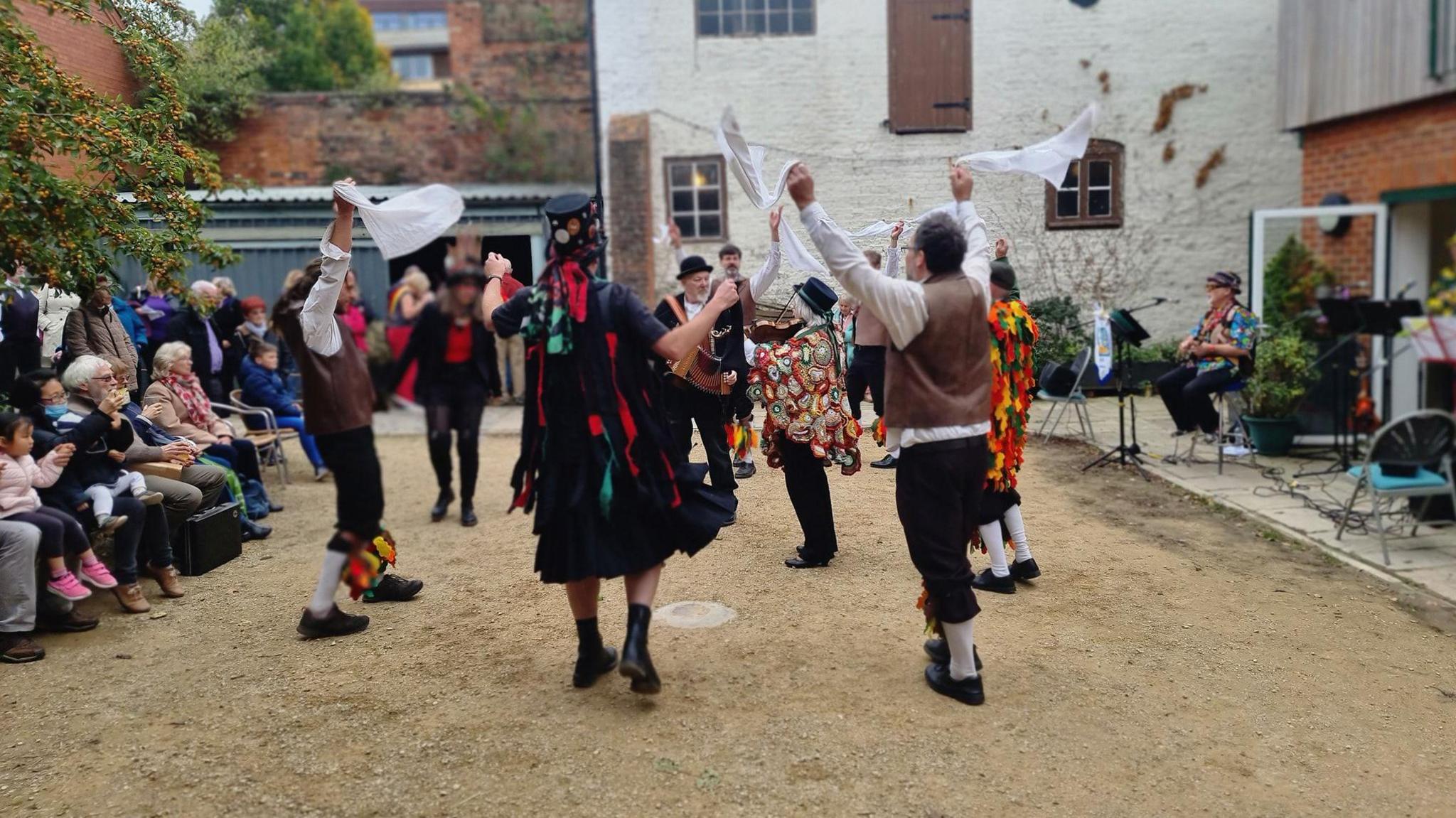 A group of Morris dancers in traditional costume are waving handkerchiefs in the air. An accordion player is in the middle of the circle of dancers wearing a top hat and black 'tatter' coat covered in ribbons.