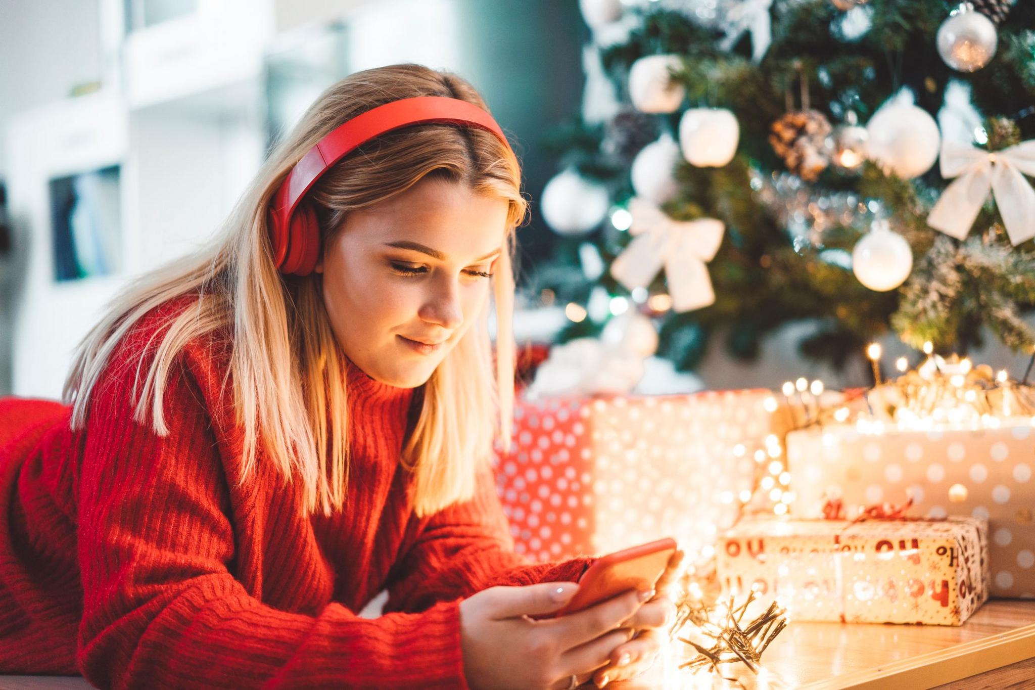 A young blonde woman in a red jumper is lying on the floor next to a Christmas tree and a pile of wrapped Christmas presents. She is looking at her mobile phone and wearing red headphones.