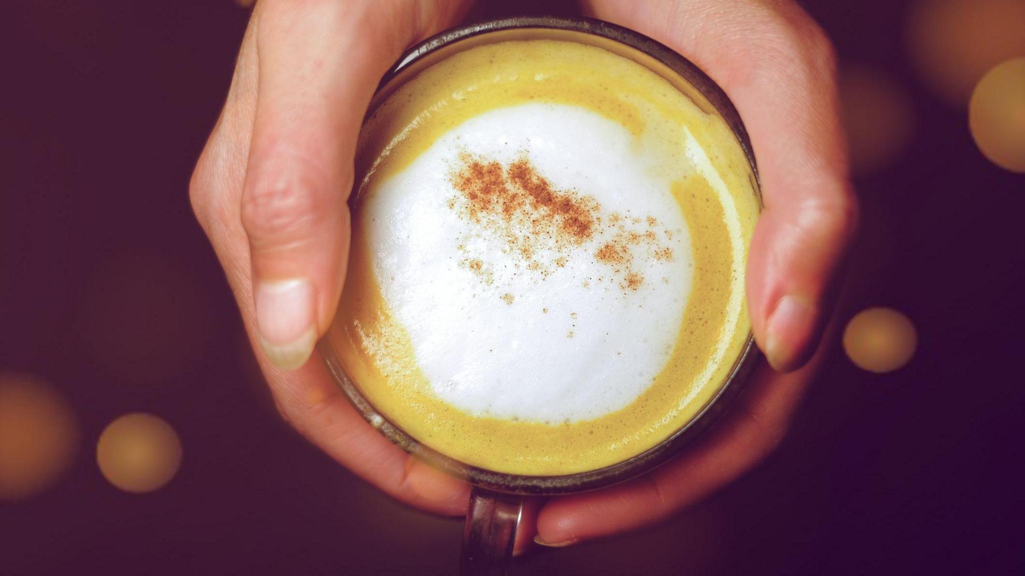 A close up photograph from above of someone's hands wrapped around a glass mug of pumpkin spice latte. It has foamed milk and a sprinkle of cinnamon on top.