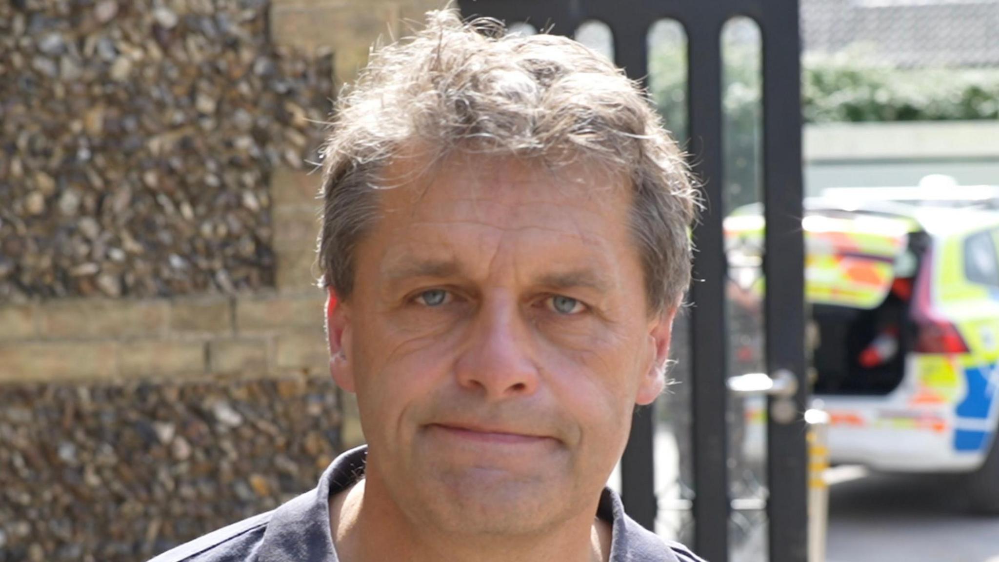 A head-and-shoulders image of a man standing in front of a wall. He has a look of resignation. He is looking straight at the camera. In the background, which is blurred, there is a police car.