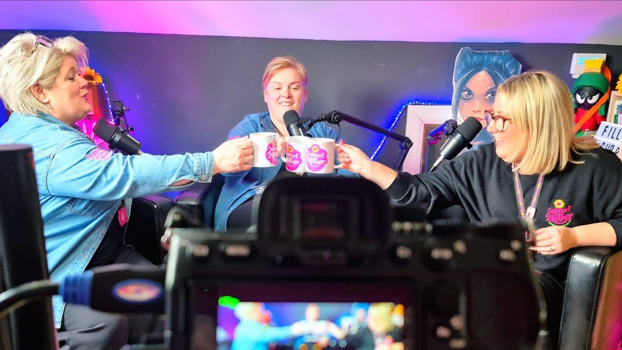 Three women smiling sitting in a triangle. They are holding mugs that have the pink logo of Support 2gether on them. The three women have blonde hair and have podcast mics in front of them. There is a black camera with the image blurred in the view finder.