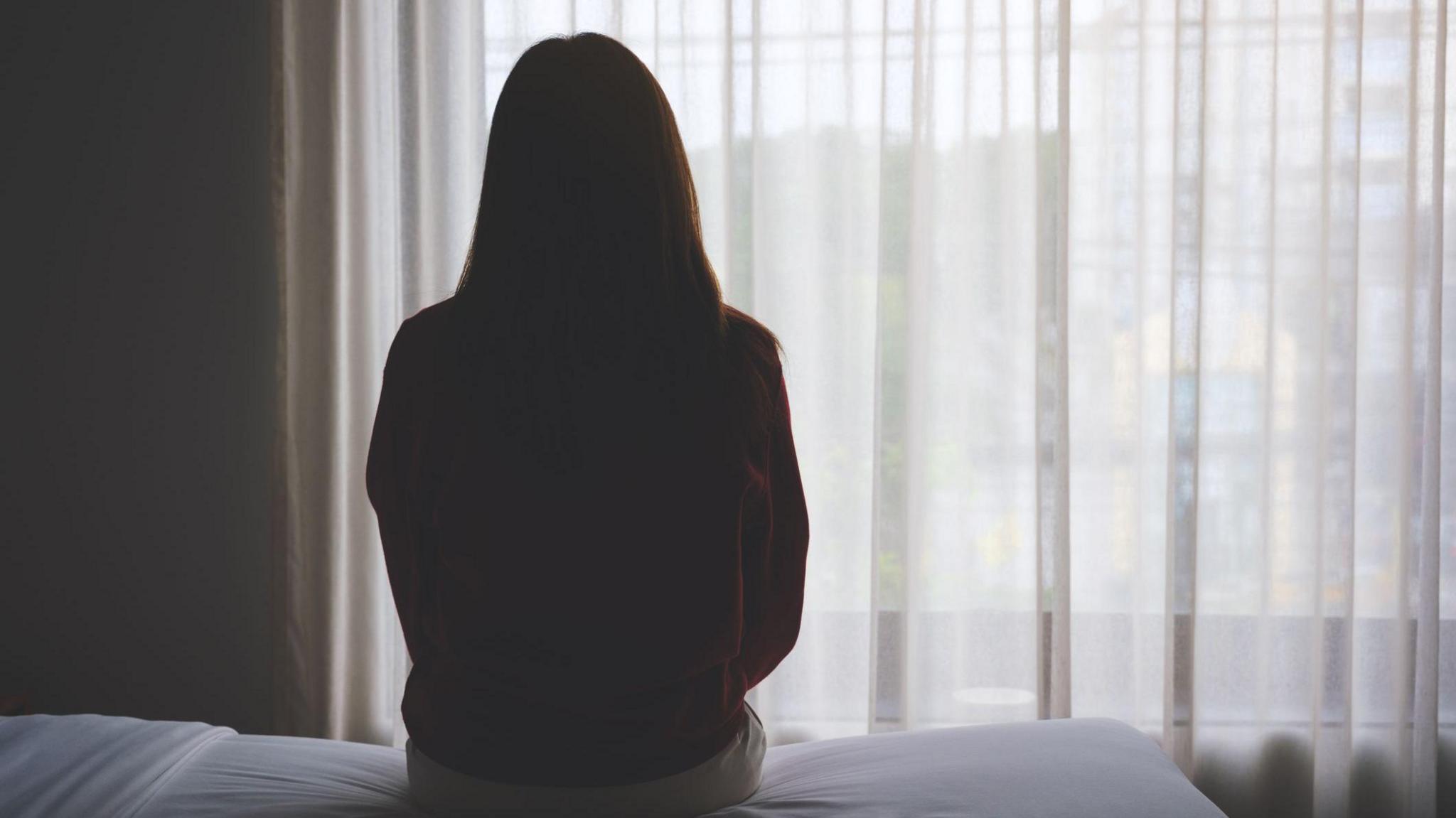 The back of a woman sitting on a bed looking out a window with curtains on it. 