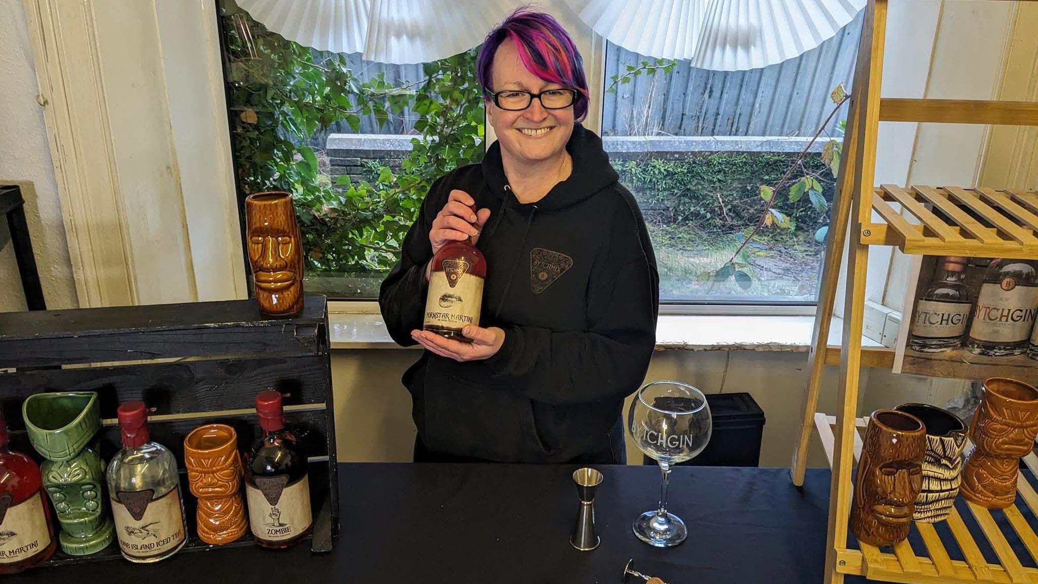 A woman stands behind a bar and in front of a window. She has purple hair with pink highlights and holds a bottle of a golden coloured drink. A spirit measure and a gin glass can be seen on the right next to a small wooden shelving unit. To the right, several other bottles of drink sit in an upright wooden box.