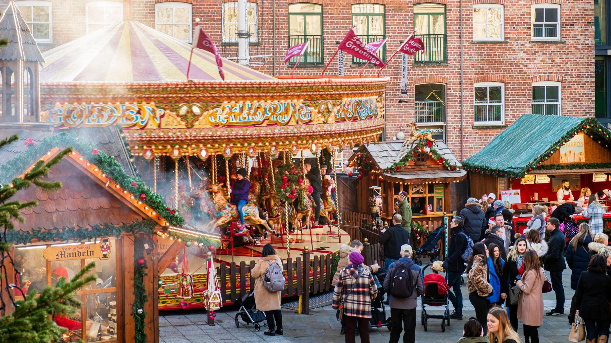 A carousel and market stalls in Leeds for the Christmas market. Many people wearing warm coats stand by the stalls watching people on the carousel