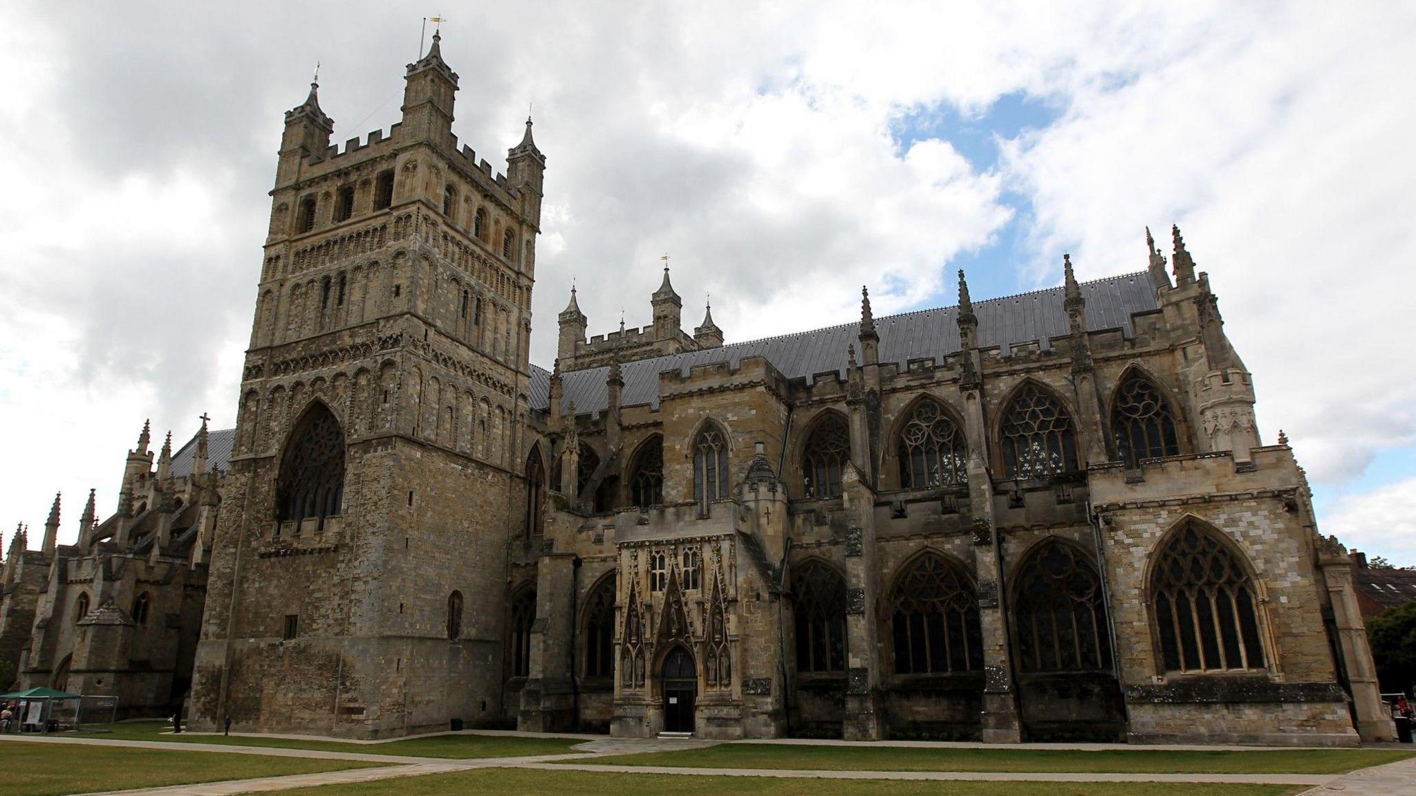 An outside shot of Exeter Cathedral. It shows all the architecture and the cathedral green. There is the main building and the tower which is at the heart of it. The weather is cloudy with a blue sky behind them.