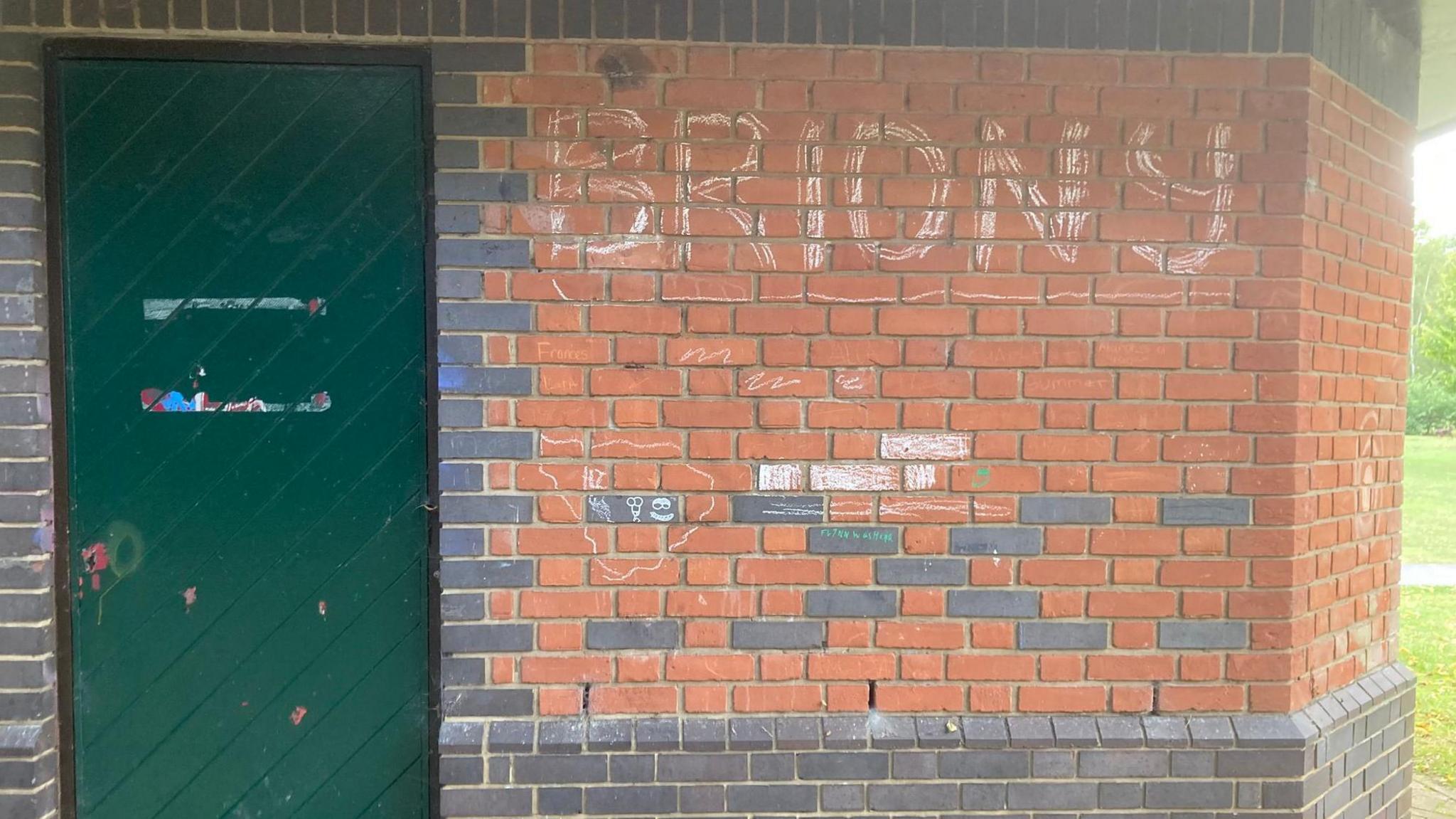 The name Briony graffitied on the side of a red-bricked wall. There is a green door to the left of the mage.