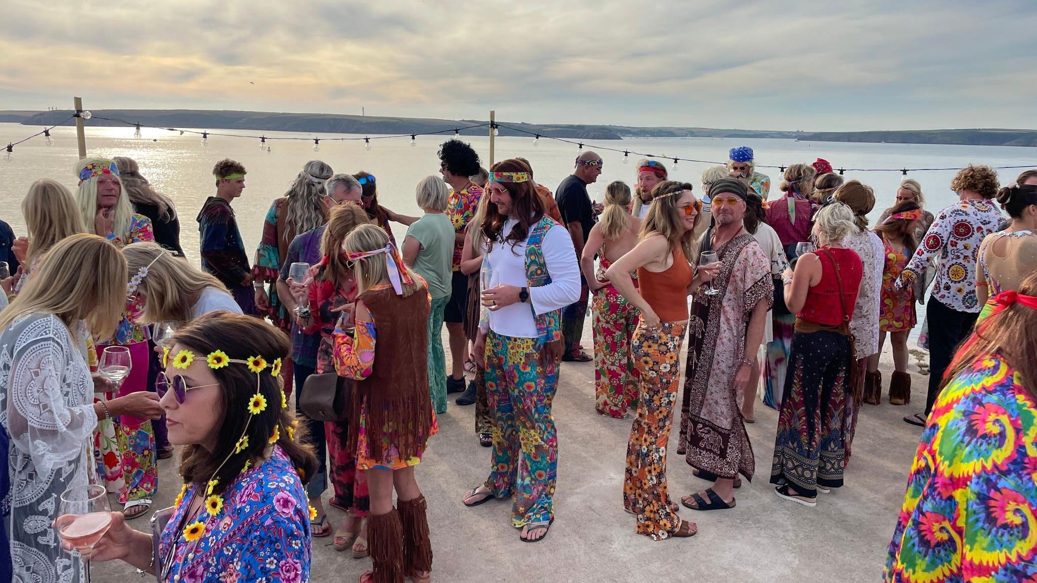 A large group of people stand on a flat concrete surface, with the sea stretching beyond then. They are all wearing colourful 60s themed fancy dress costumes, with many wearing different coloured round sunglasses, long wigs, and headbands.  