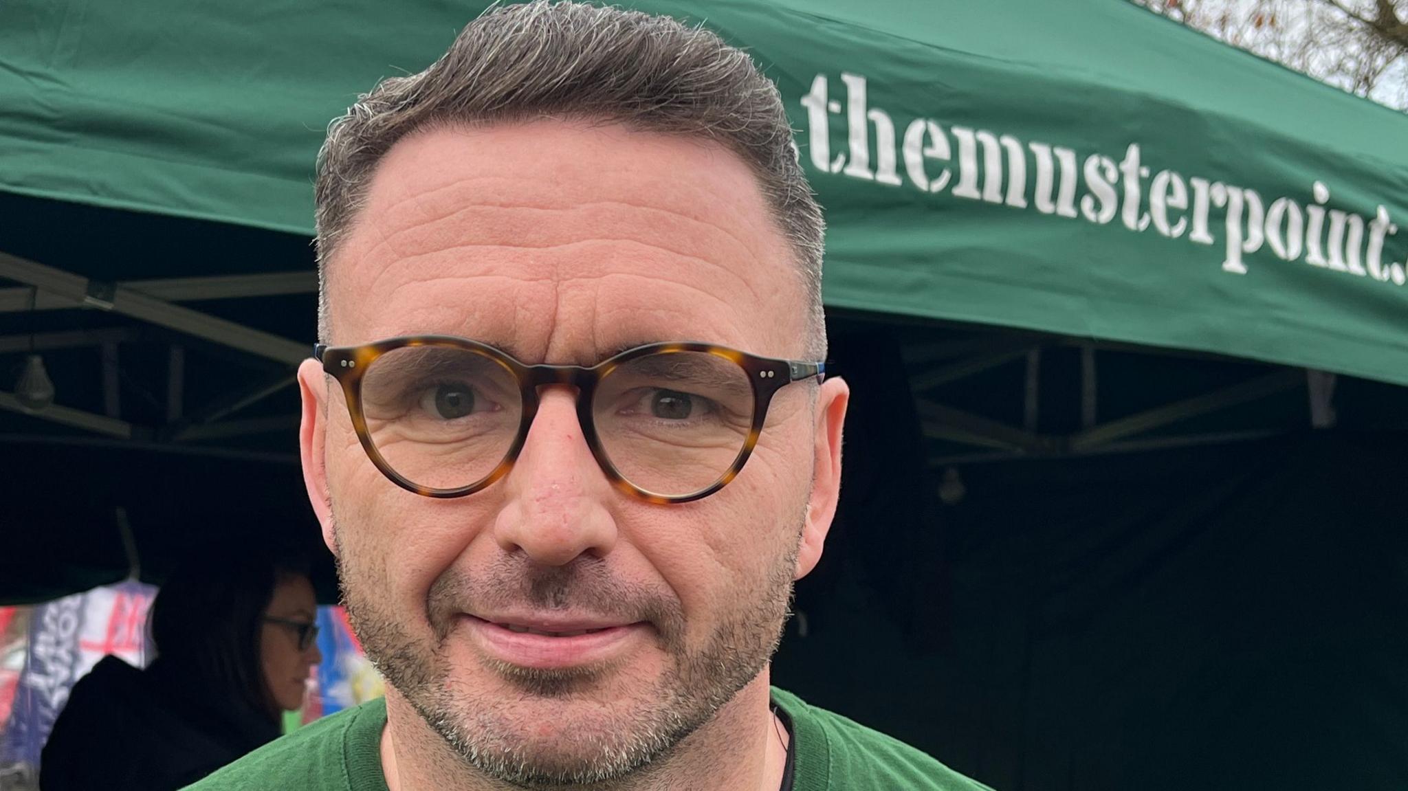A man wearing glasses and a green t shirt smiles slightly as he looks into the camera, standing in front of a green gazebo bearing the words the muster point