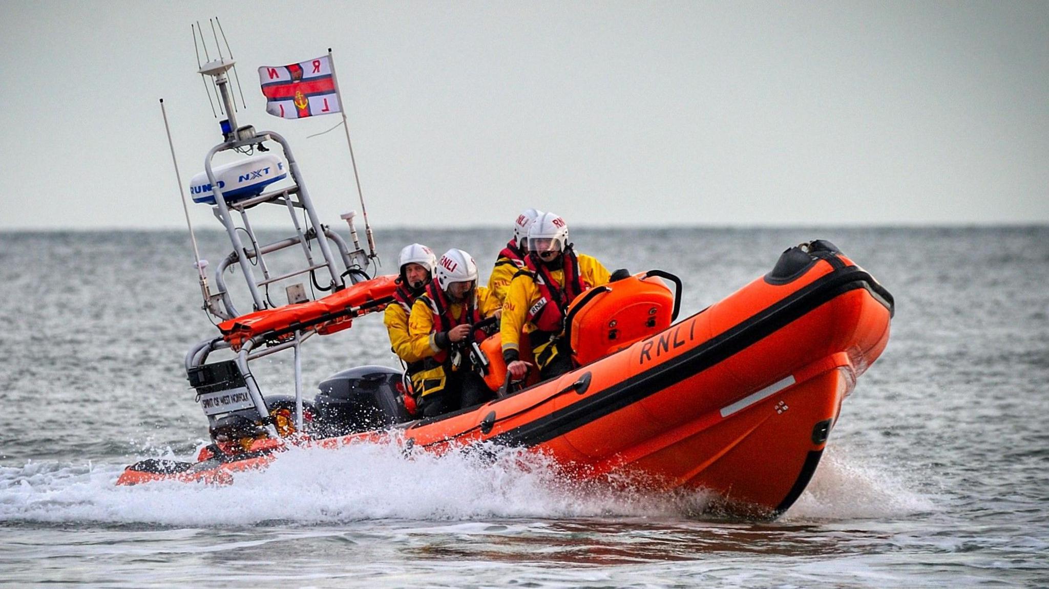 Small orange RNLI lifeboat, with four crew inside wearing white helmets, life vests and yellow coats, moving in the water and creating splash.