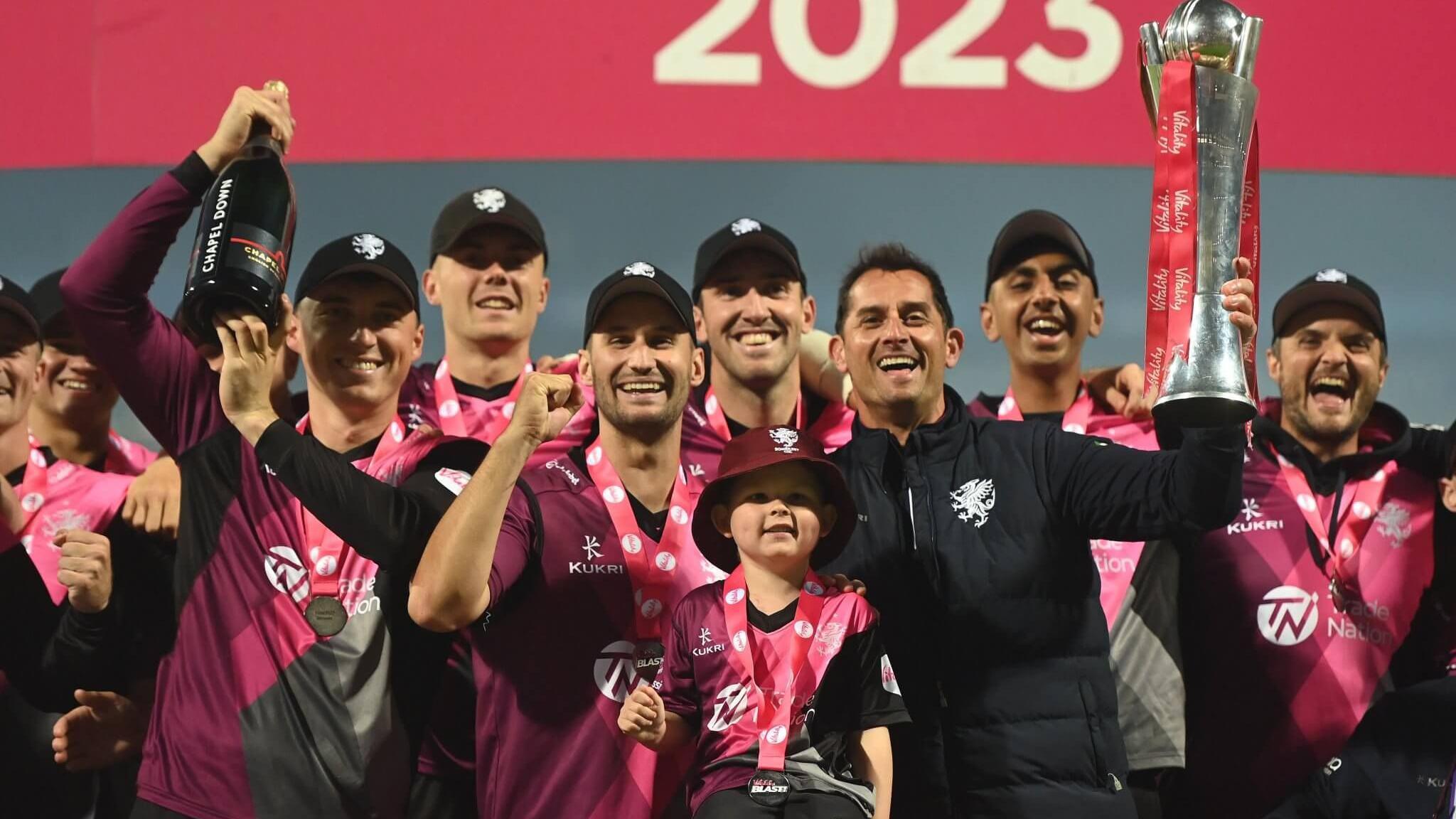 Bodhi stands in front of a cricket team, all wearing pink and black shirts. Bodhi is wearing a burgundy hat and is right next to a smiling man holding a large silver trophy. The rest of the team are smiling widely too and one man is holding up a large bottle of bubbly.