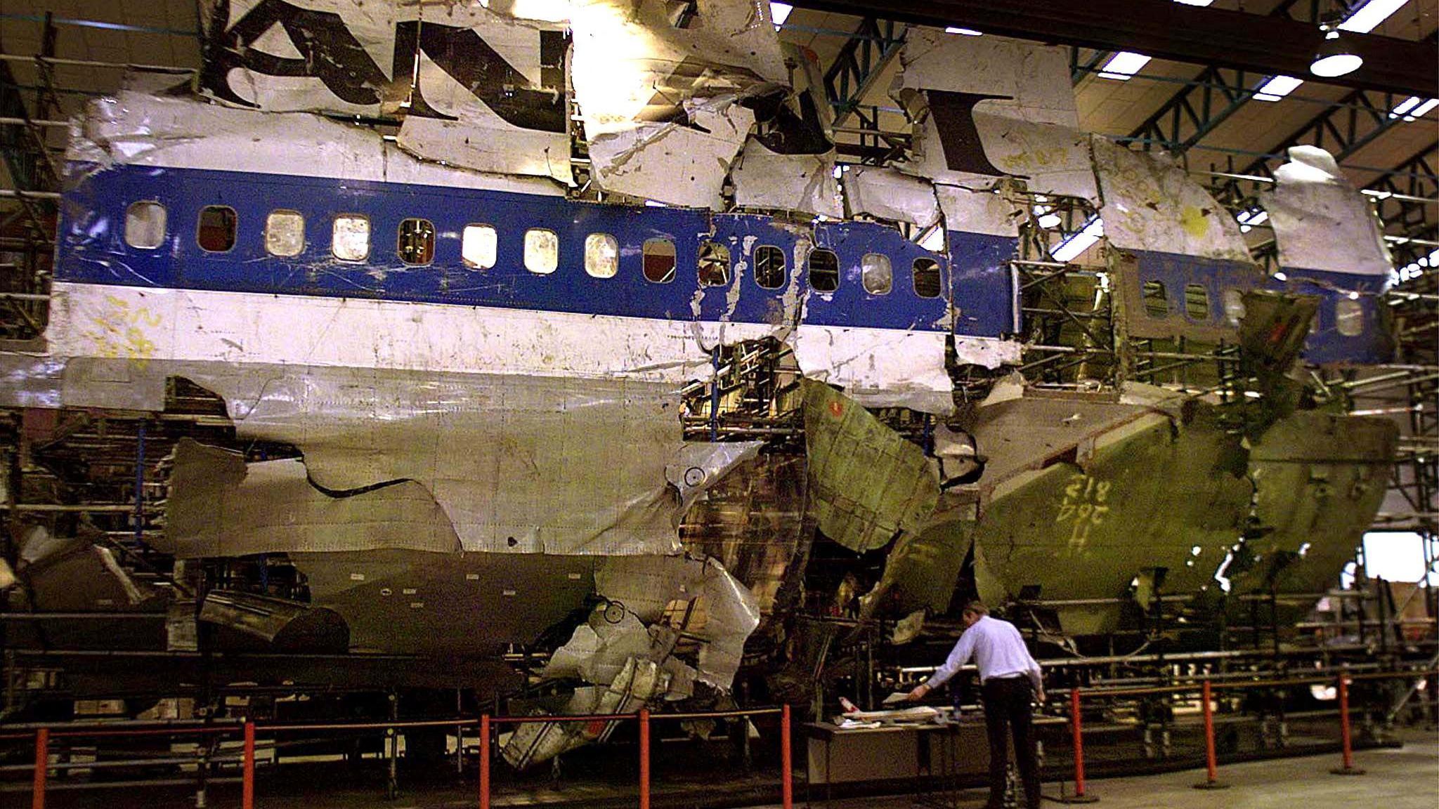 The wreckage of the plane in a warehouse. It is white with a blue stripe and black lettering, and it has missing and damaged sections. A man in a white shirt and black trousers is standing in front of the wreckage.