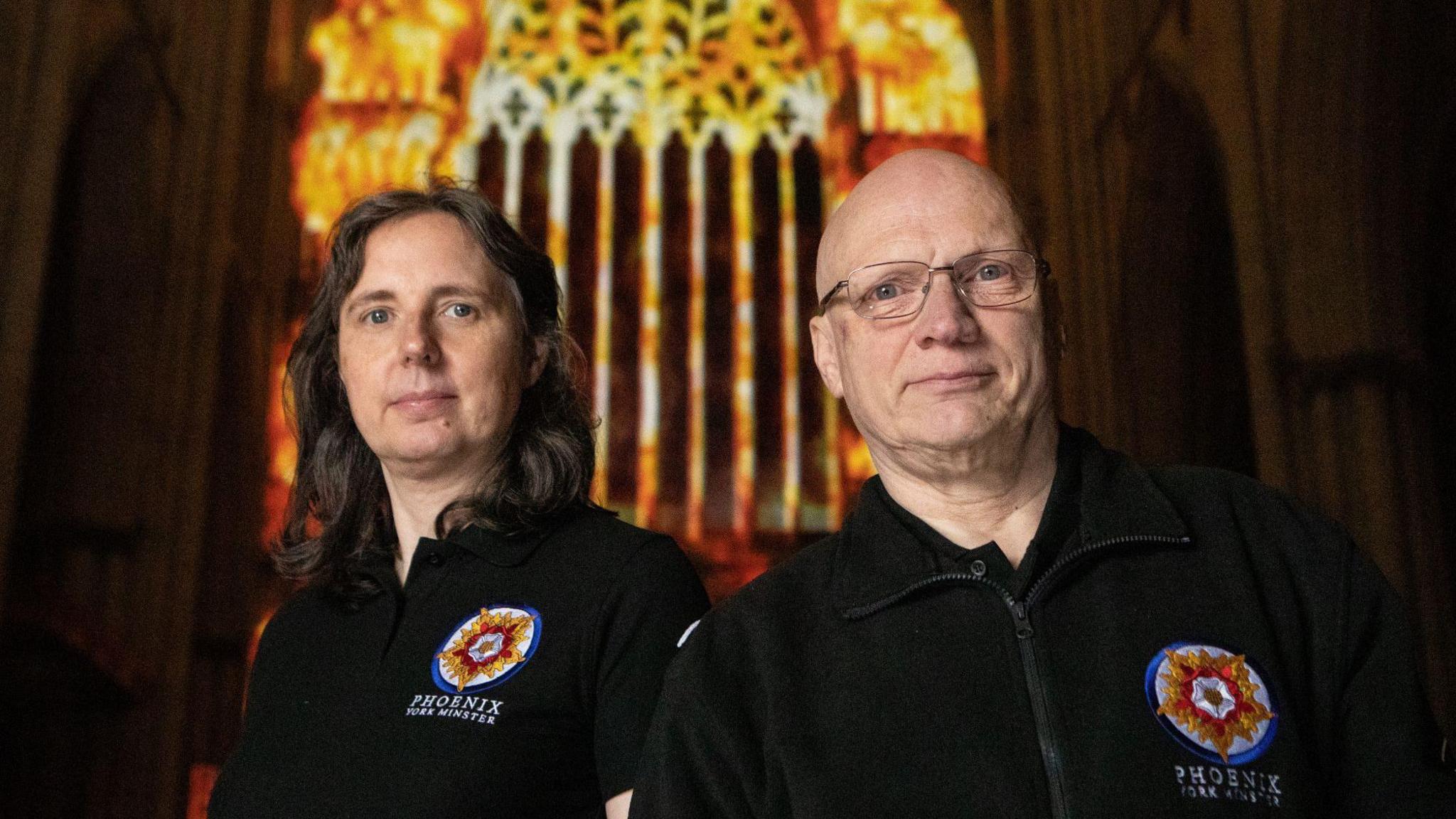 Artists Karen Monid and Ross Ashton stand in front of the light installation in York Minster. Both are wearing clothes branded with "Phoenix York Minster". Ross has glasses and a shaved head, with Karen having long dark hair. 