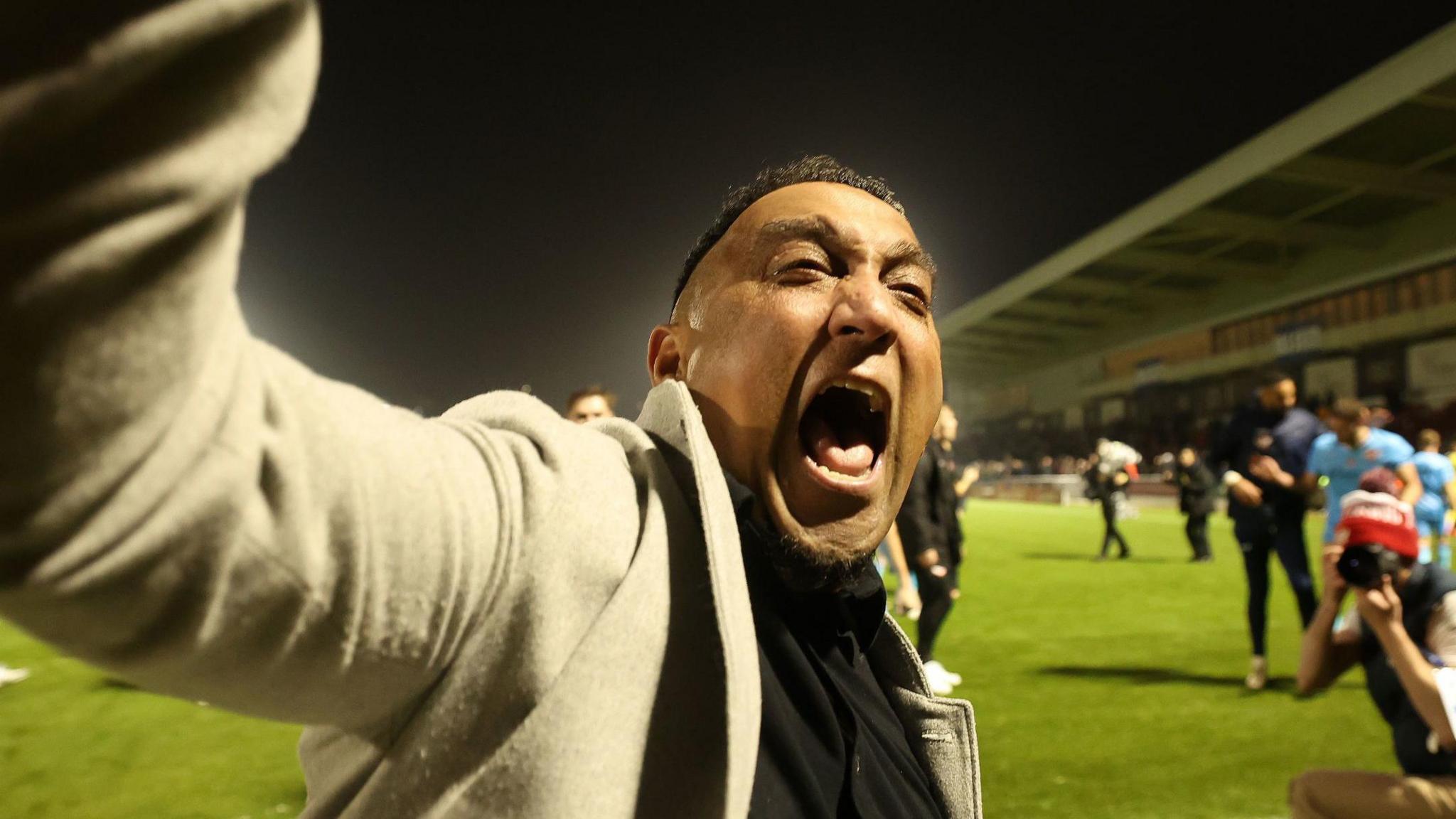 George Akhtar shouting in celebration after Kettering Town's win over Northampton. He is on the pitch standing in front of away fans who are out of view. Akhtar has his right arm lifted high in celebration.