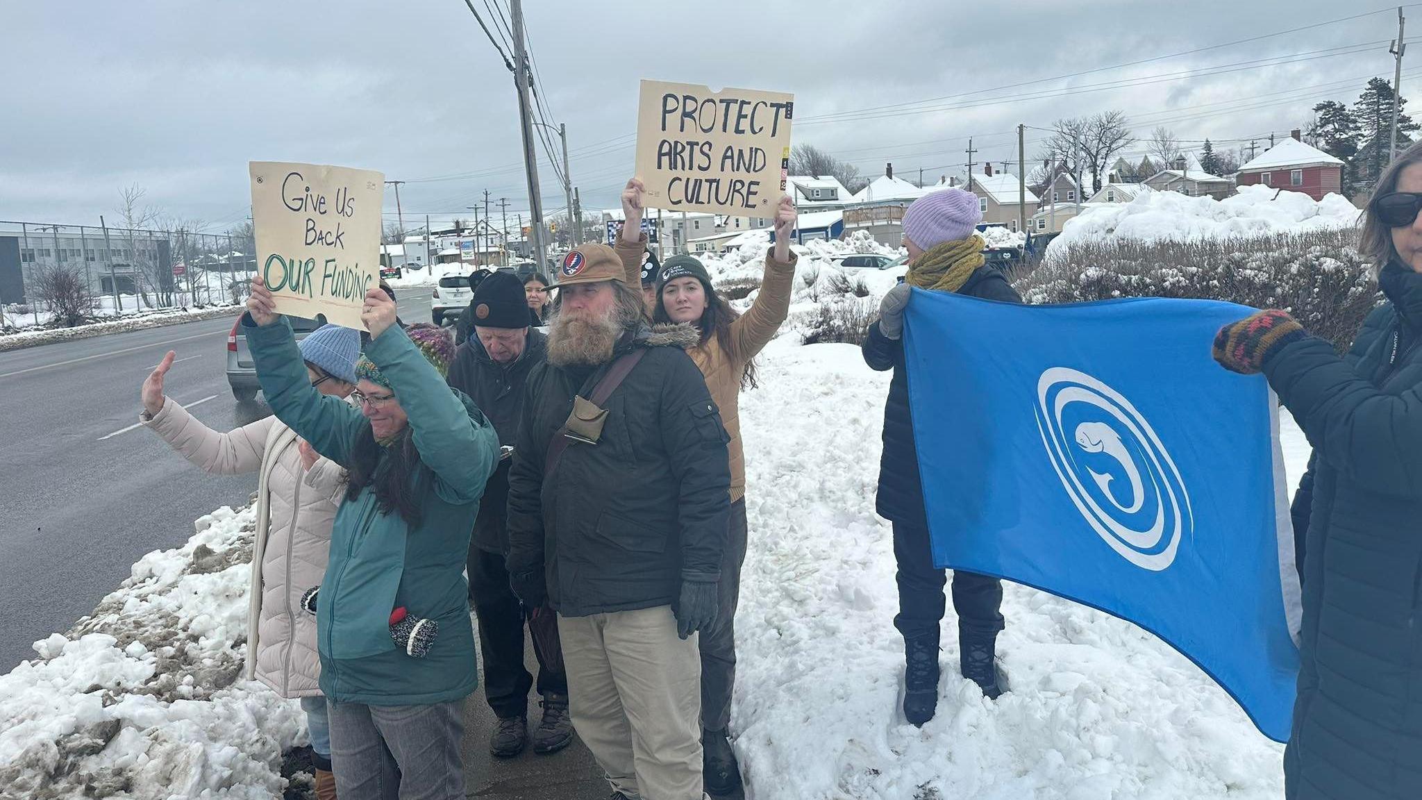 Daoine nan seasamh san t-sneachd an cois rathaid le bratach Comhairle na Gàidhlig 's iad a' togail fianais ris an trafaig
