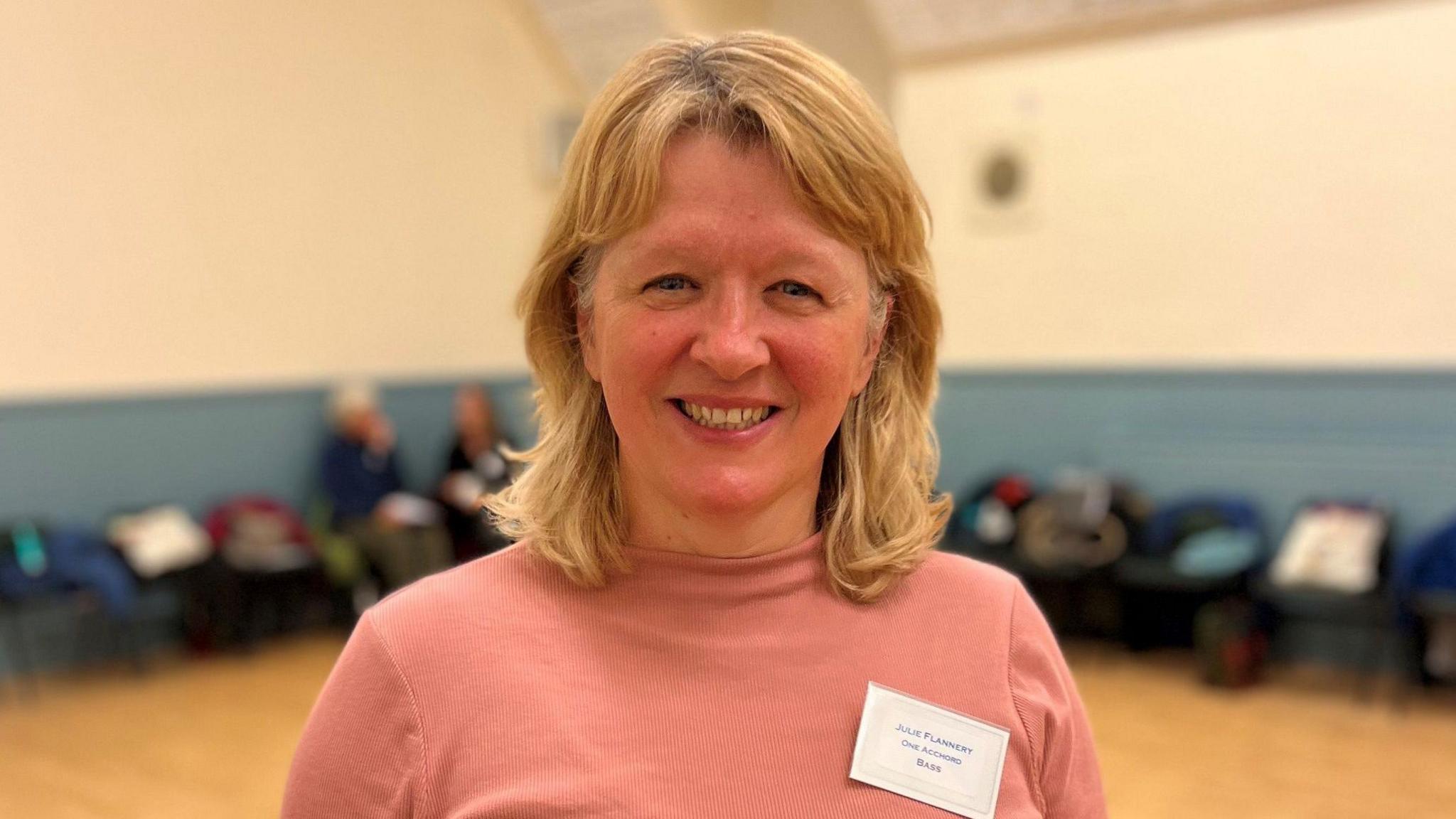 A blonde haired woman with a pink top is smiling at the camera