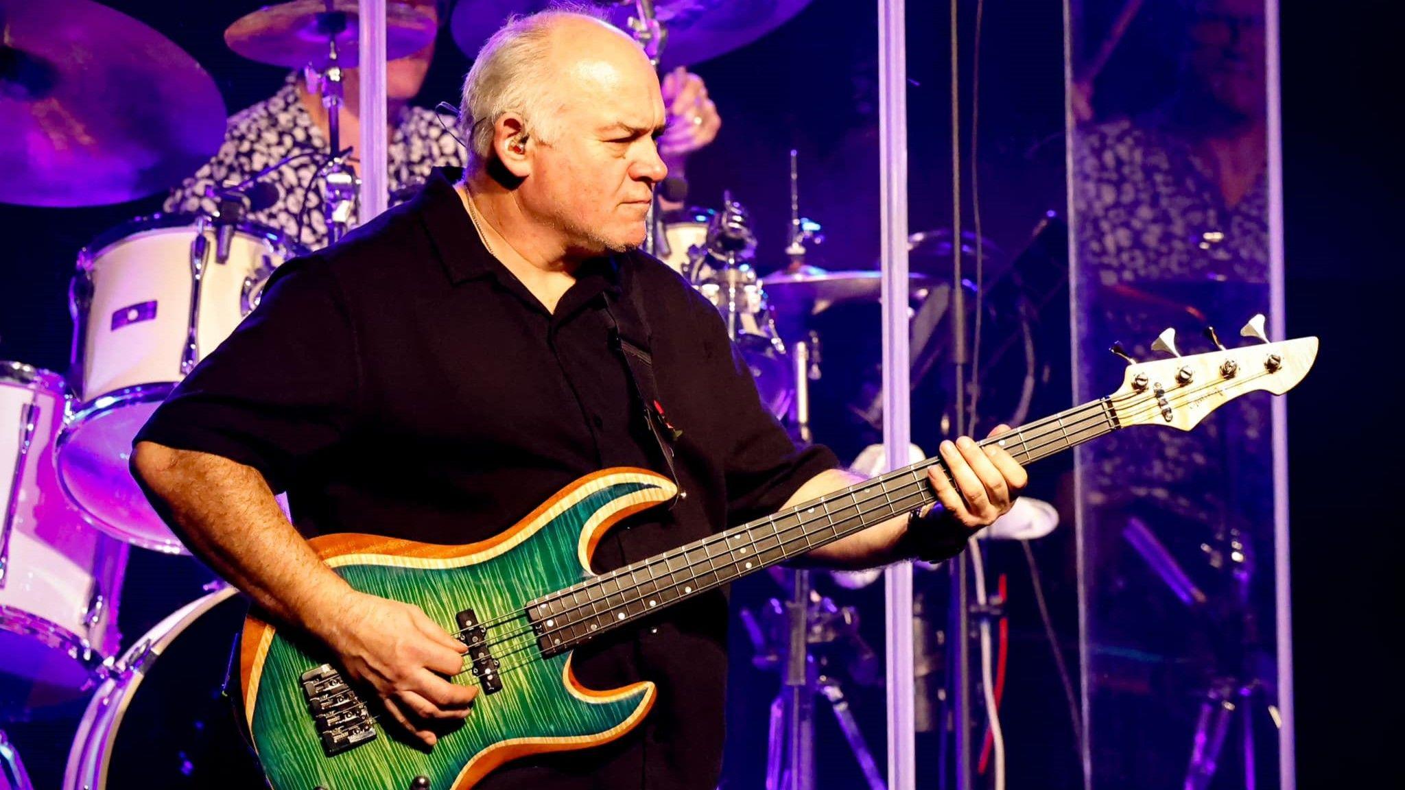 Nick Smith playing a green and orange bass guitar on stage. He is balding with grey hair and is wearing a black polo shirt. A drummer can be seen behind him.