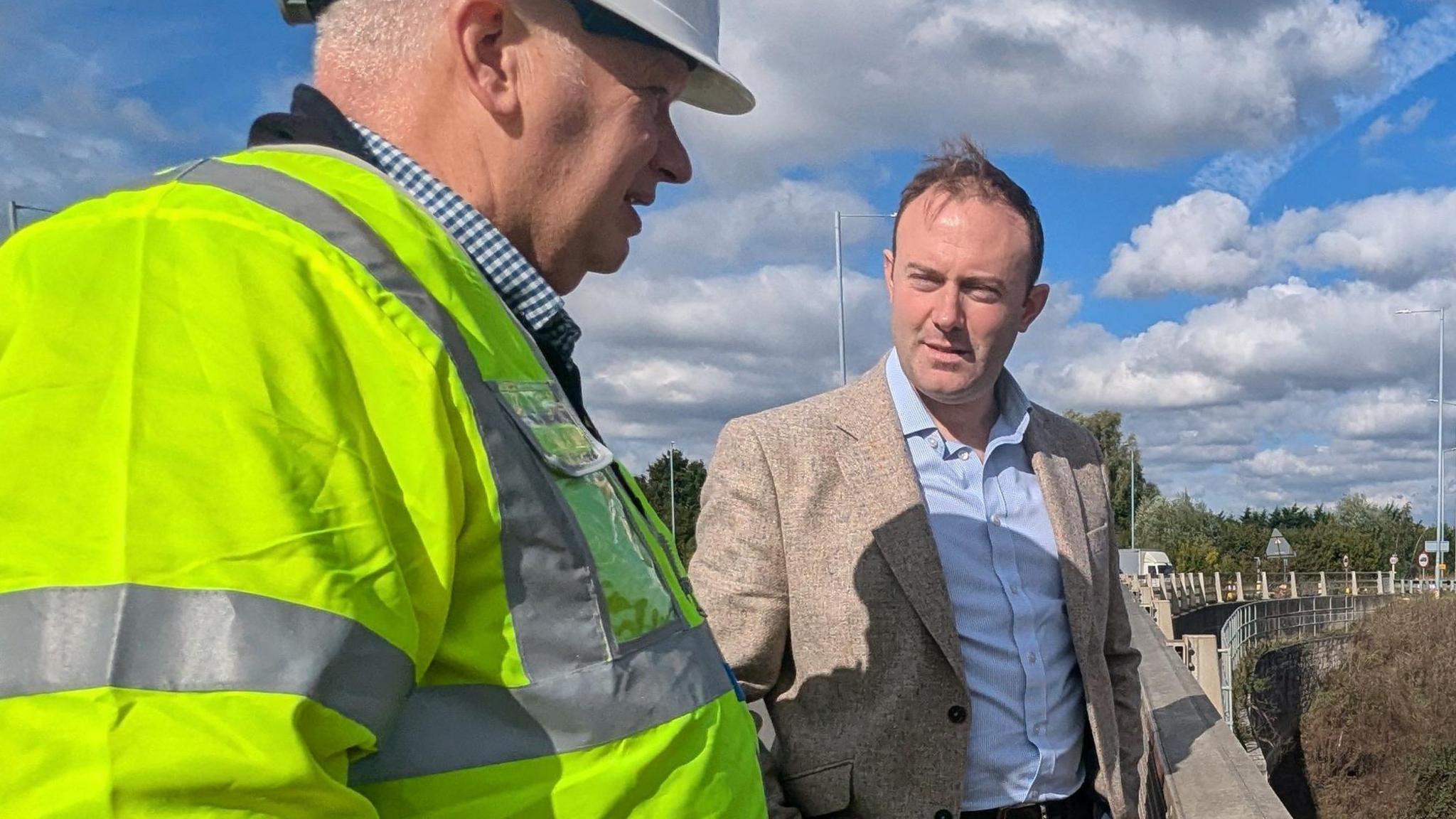 Blake Stephenson and another man standing outside on what appears to be a road overpass. Stephenson is wearing a light coloured blazer and blue shirt. Another unidentified man is wearing a high visibility jacket and a white hard hat. The collar of this man's blue checked shirt is just visible. The two men appear to be in conversation with one another. The road can be seen disappearing off into the background. It is a bright, sunny moment, though there are puffy white clouds in the sky.