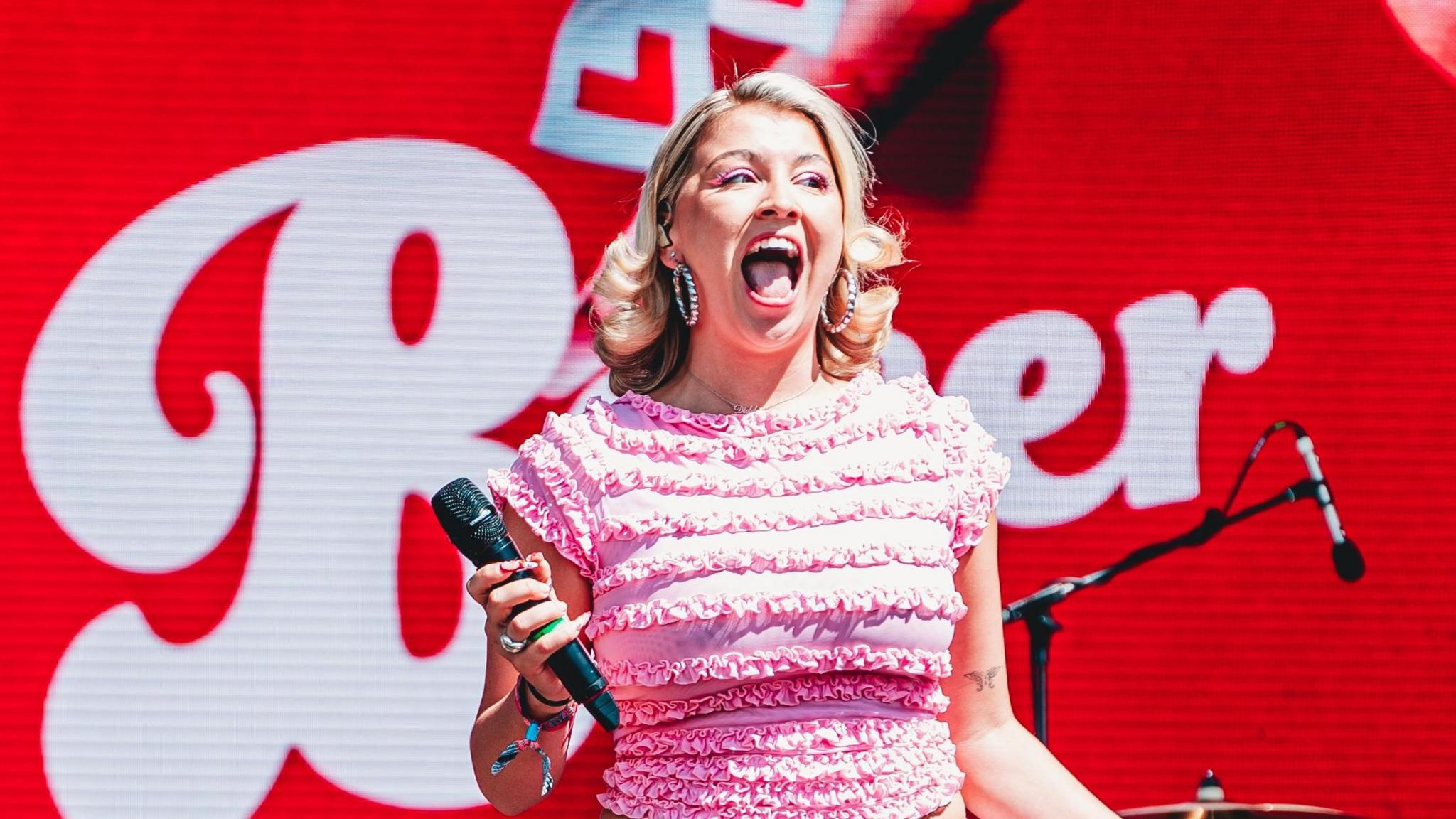 Caity Baser performing on stage. She wears a frilly pink top, hoop earrings and red lipstick, her mouth open wide as she seems to scream in excitement towards the audience. The staging behind her is red, with 'L' plates and her name in a white font. 