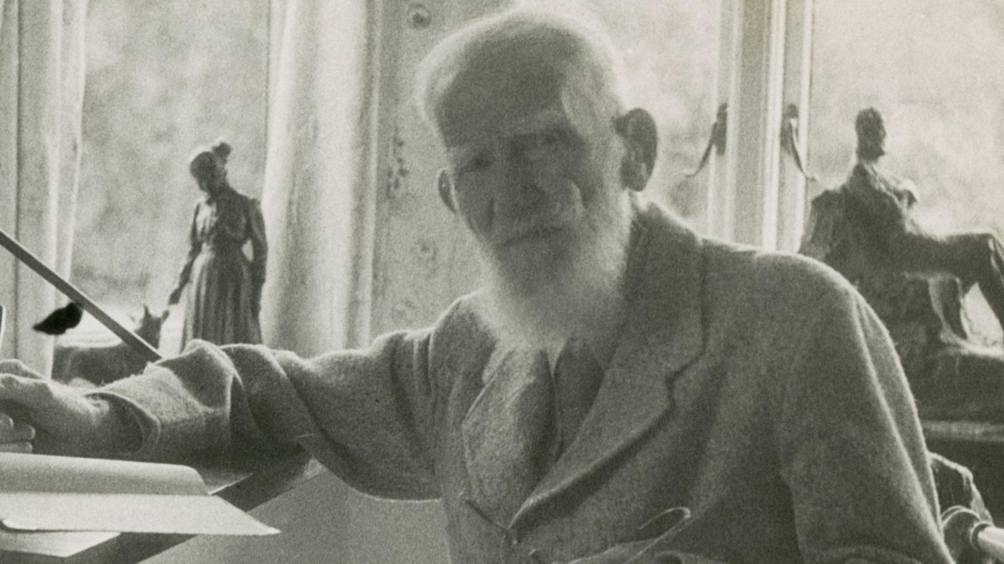 A grainy old black-and-white photo of George Bernard Shaw. He is dressed smartly and has a long white beard. He is sitting at a writing desk holding a pen.