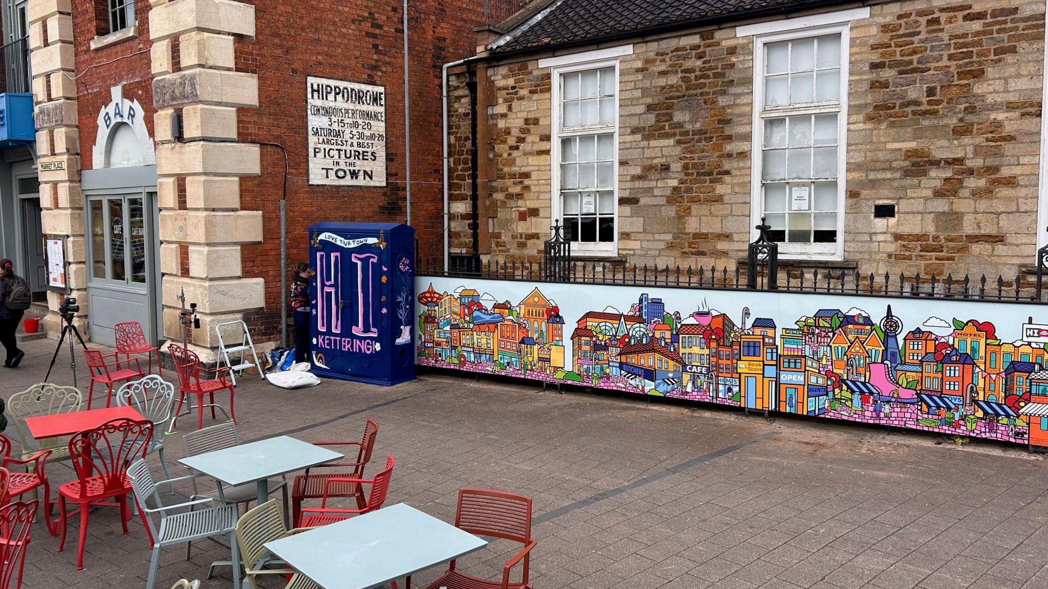 A purple electricity box is painted purple with "Hi Kettering" written on it. Next to it is a colourful mural of different coloured buildings against railings.