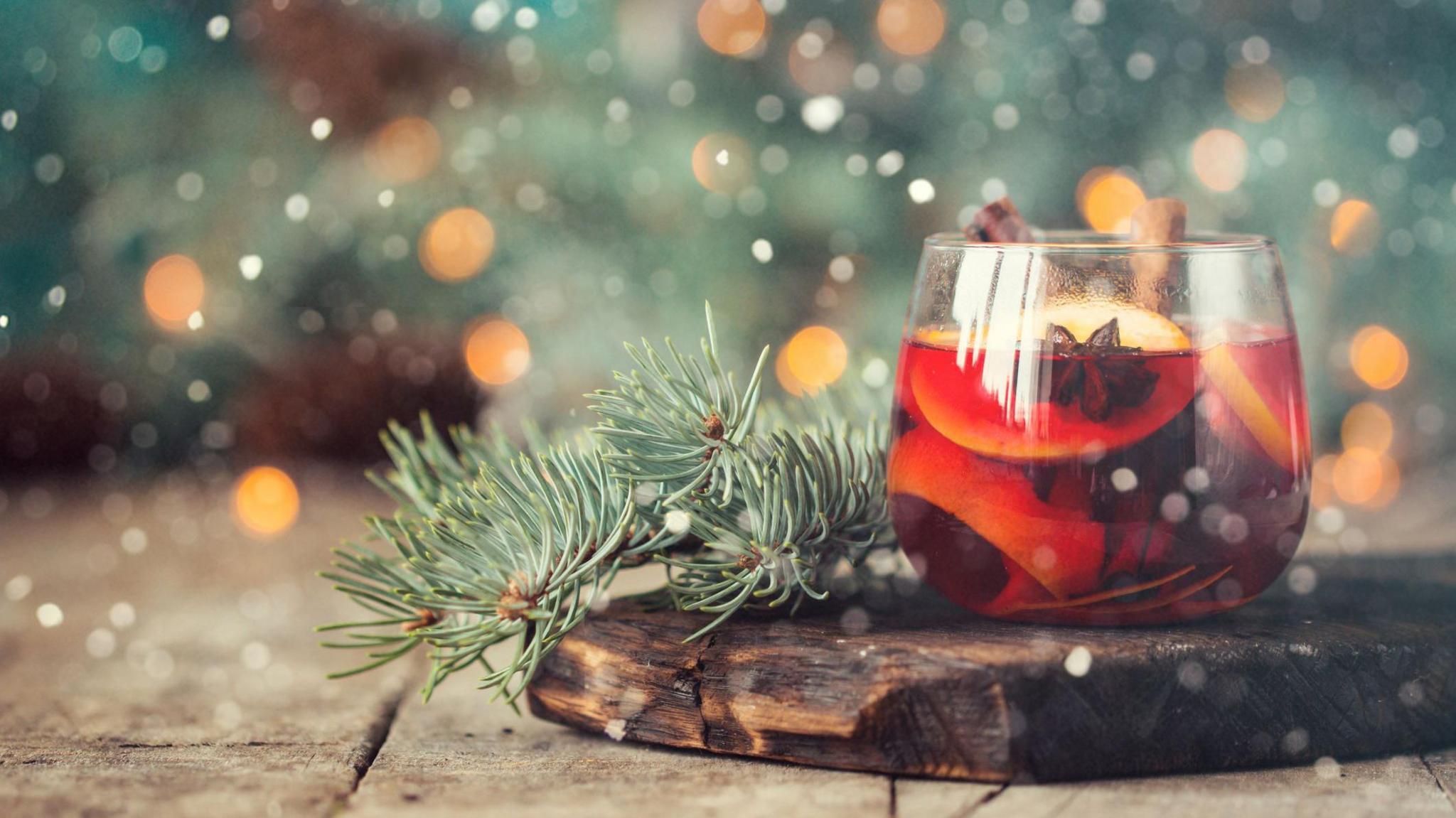 A glass tumbler filled with a red liquid and slices of orange, meant to resemble mulled win, stands on a wooden board with two green foliage fronds and a sparkly background