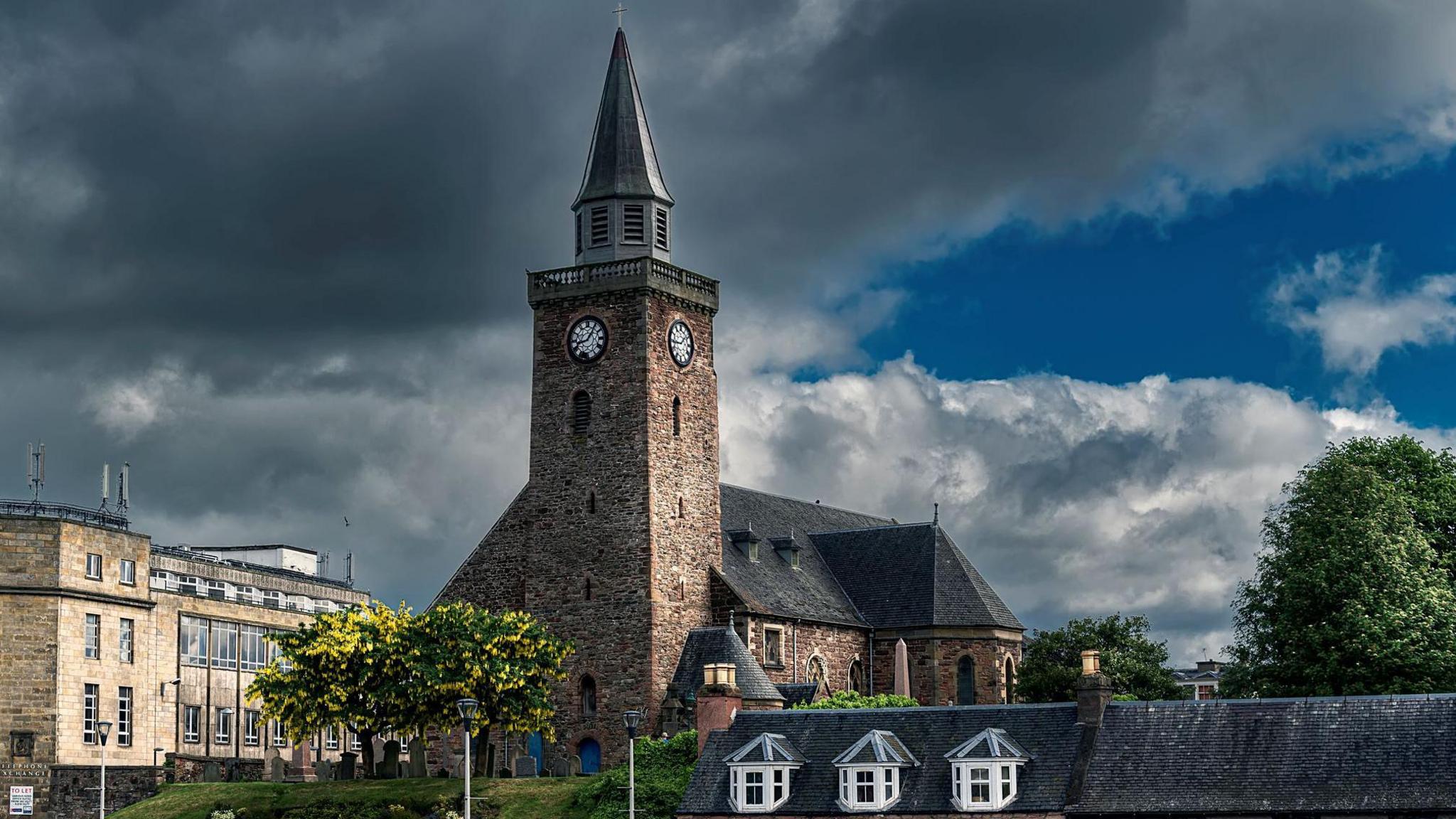 The church's steeple catches sunlight but the sky behind it is dark with cloud. Next to the church is Inverness' BT building and on a street below a row of small houses.