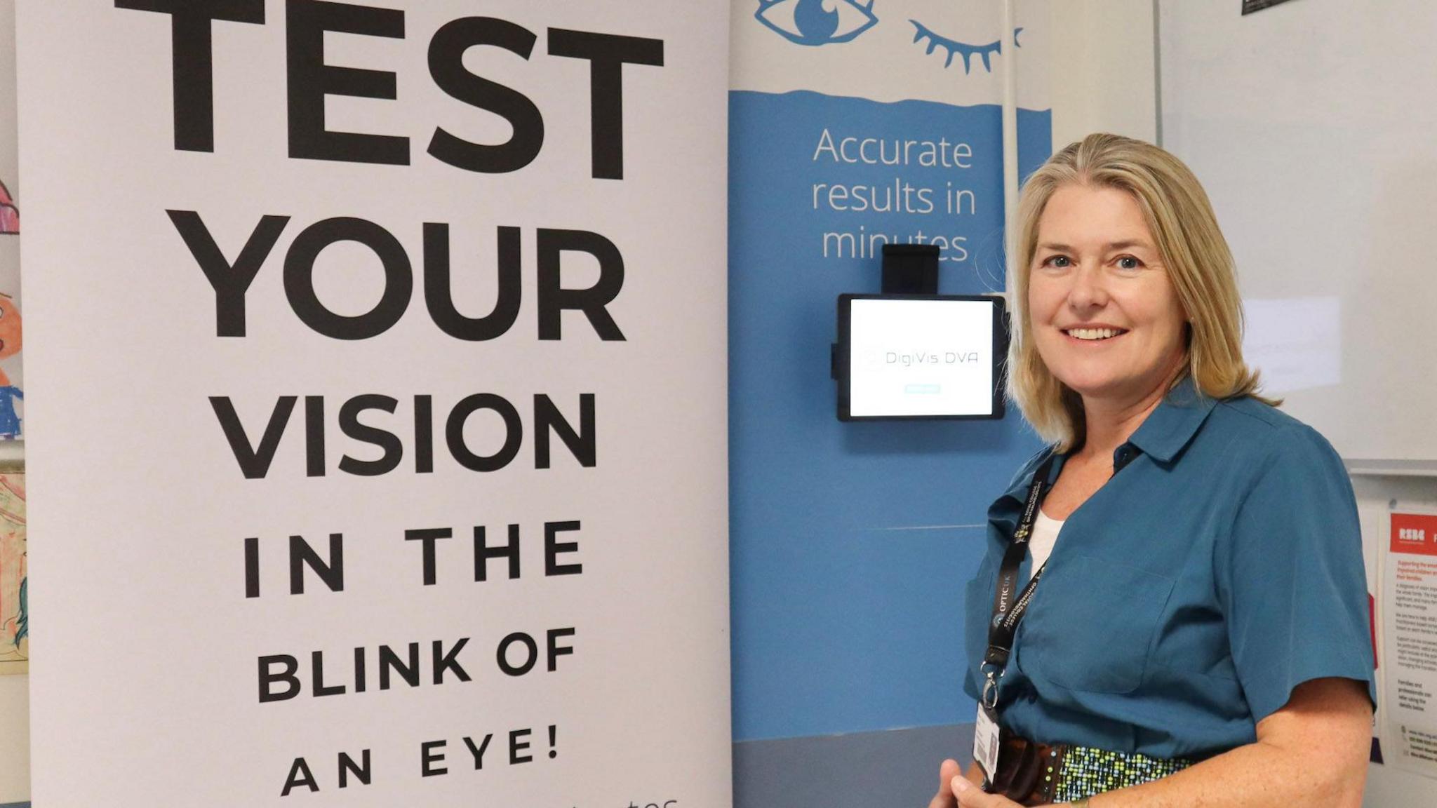Dr Louise Allen is standing in front of a large sign that says "test your vision in the blink of an eye". There is a tablet on the wall and another sign that says "accurate results in minutes". Dr Allen has shoulder length fair hair and is smiling at the camera. She is wearing a blue blouse and has a name tag around her neck