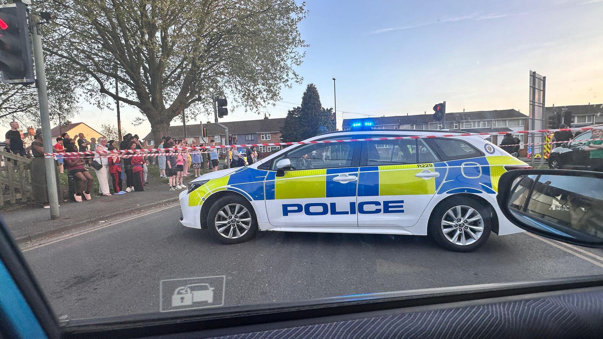 A cordon in place, tied to two traffic light poles. a Police car is parked infront of the area with onlookers at the side of the road looking into the distance.