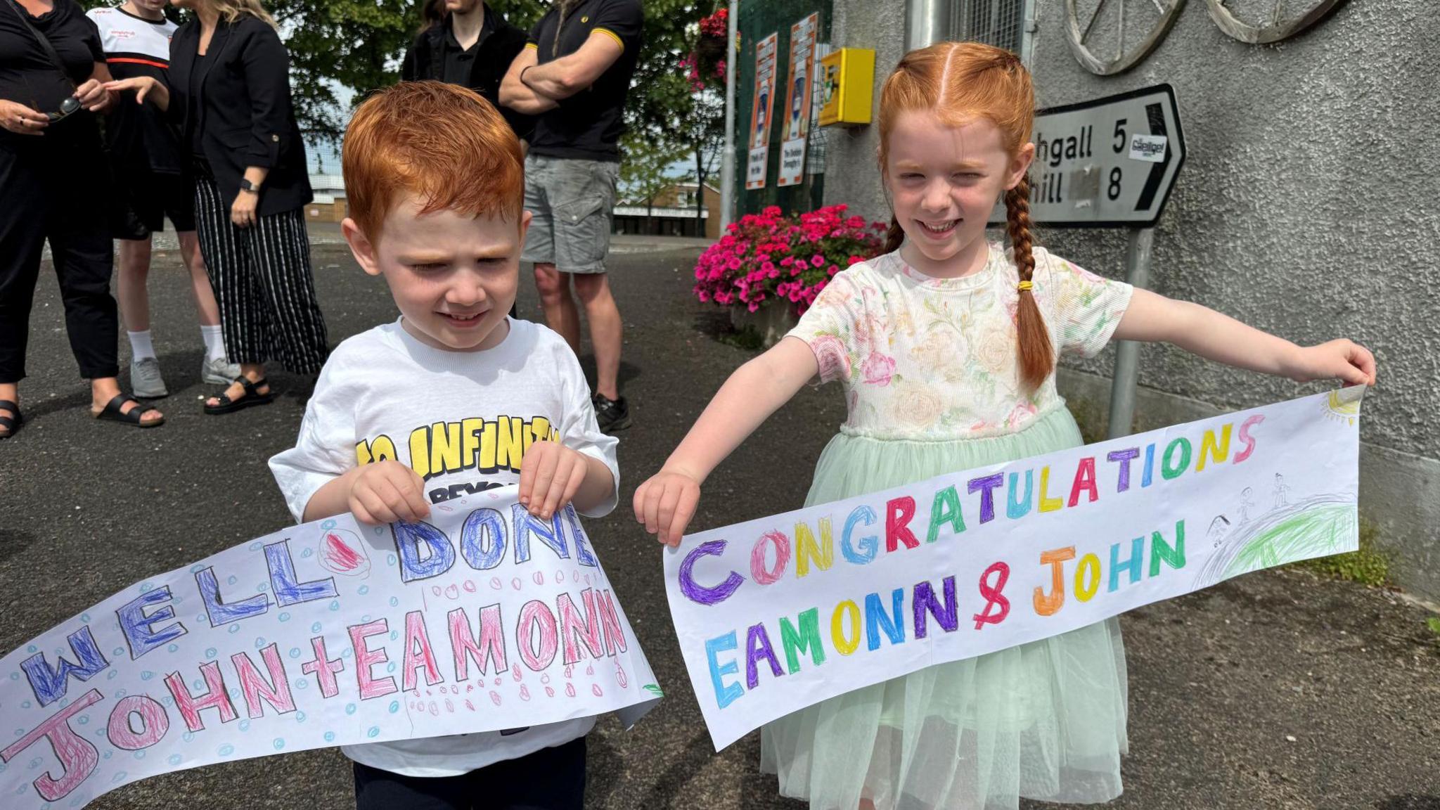 Two red haired chidren, a boy and a girl, hold colourful handmade banners, their greeting, all in uppercase letters, filled in bold colours. Behind them adults stand around talking. 