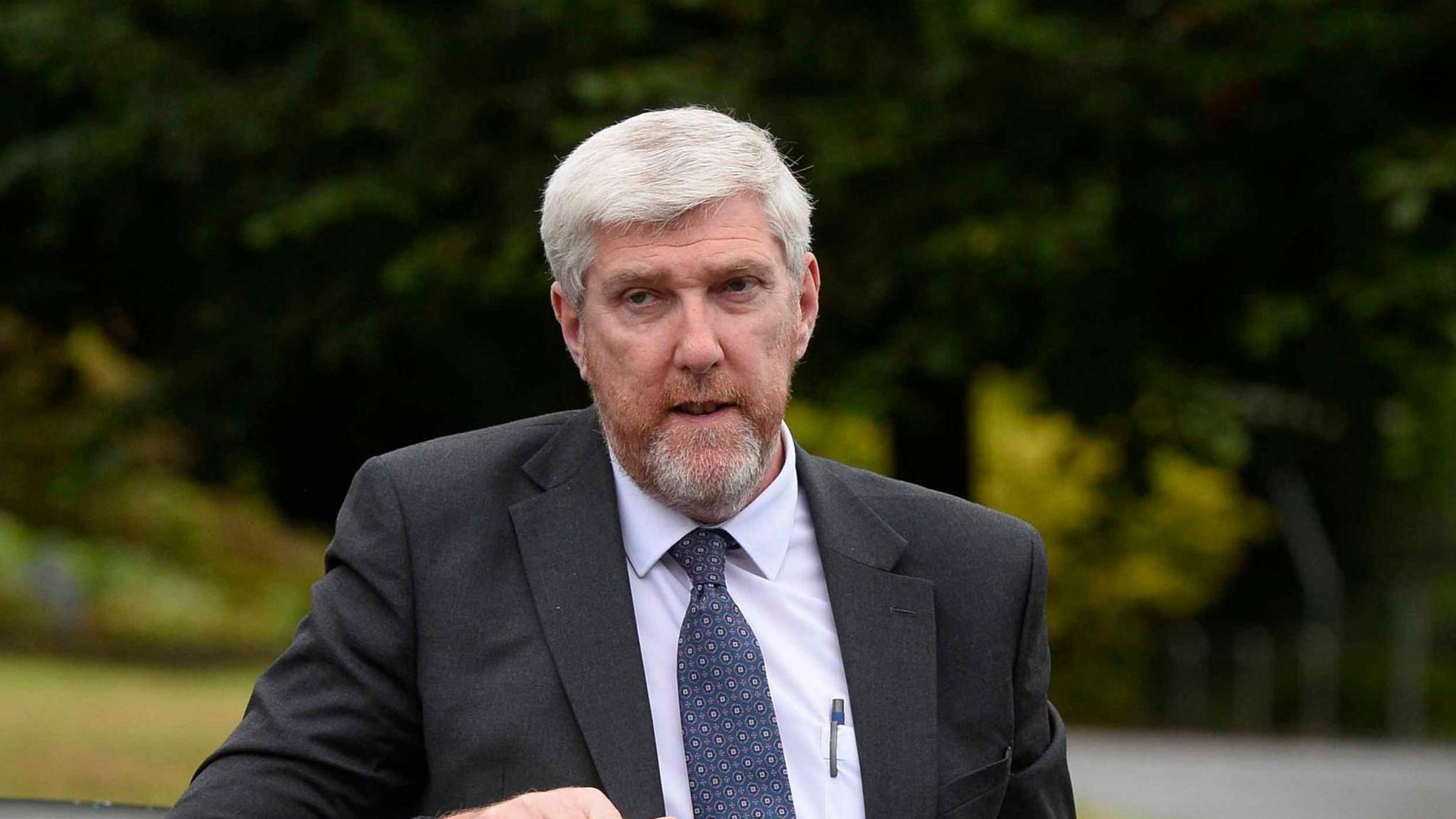 John O'Dowd dressed in a grey suit with a grey beard and a pen in his chest pocket. He has white hair and a white beard and is stood in front of trees.