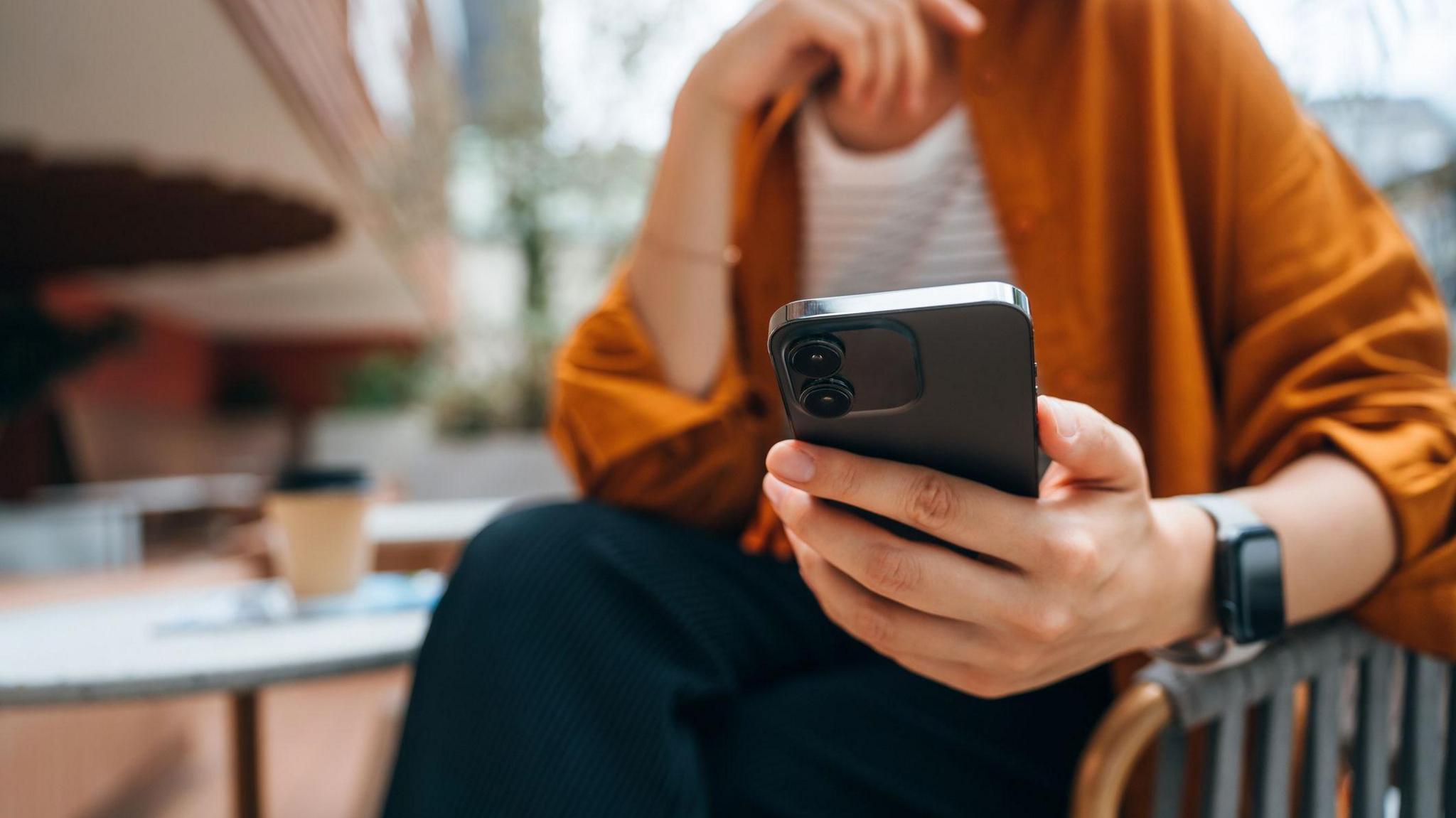 A person in an orange shirt sitting on a chair holding a phone. Their other hand rests on their chin. A blurred background shows a table with a coffee cup.