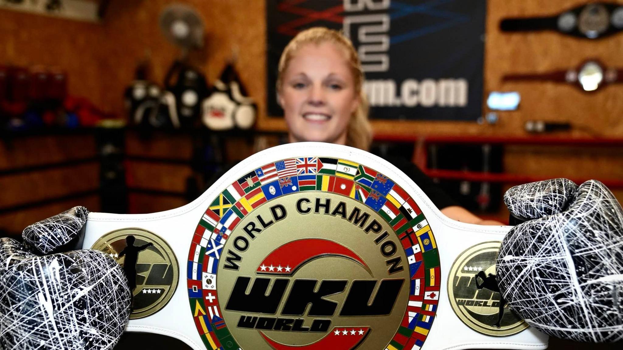 Turner is seen holding her WKU K1 World Champion belt. She is smiling while holding the white belt with a gold plate detailing the title.