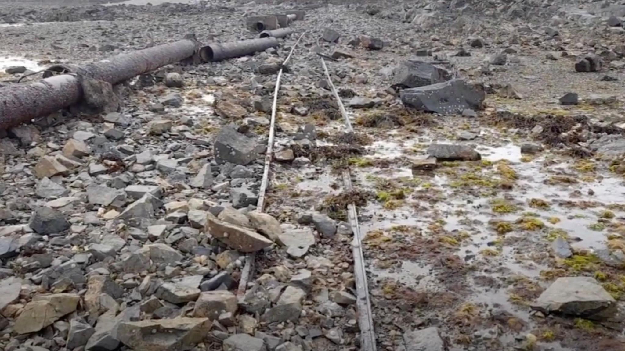 Corroded rail tracks on a rocky beach next to a rusted and broken iron outlet pipe at low tide.